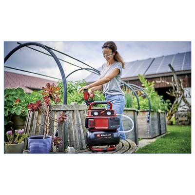 Woman operating a red garden pump beside raised beds in a backyard greenhouse garden, with solar panels visible on the roof.<br>