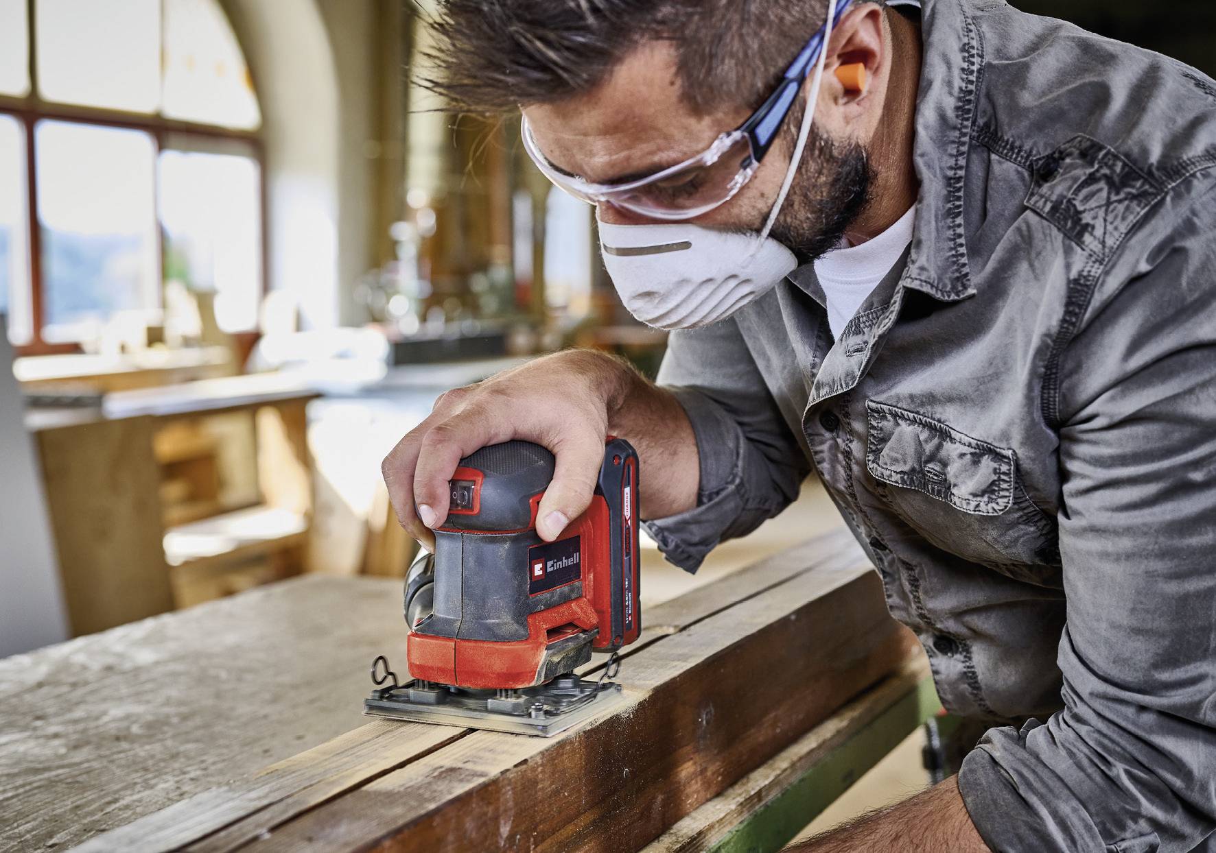 A craftsman is sanding wood with an electric sander in a workshop. He is wearing safety glasses and a mask.