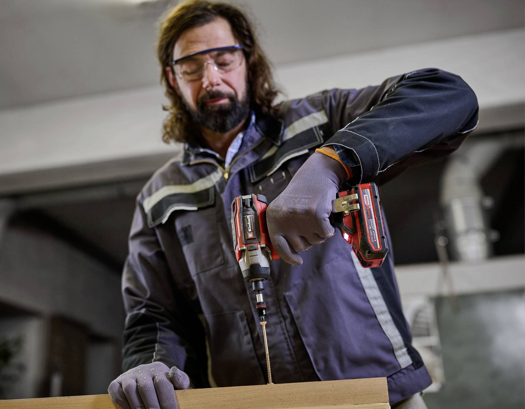 A man wearing safety glasses and gloves is drilling a hole in a piece of wood with an electric drill in a workshop.