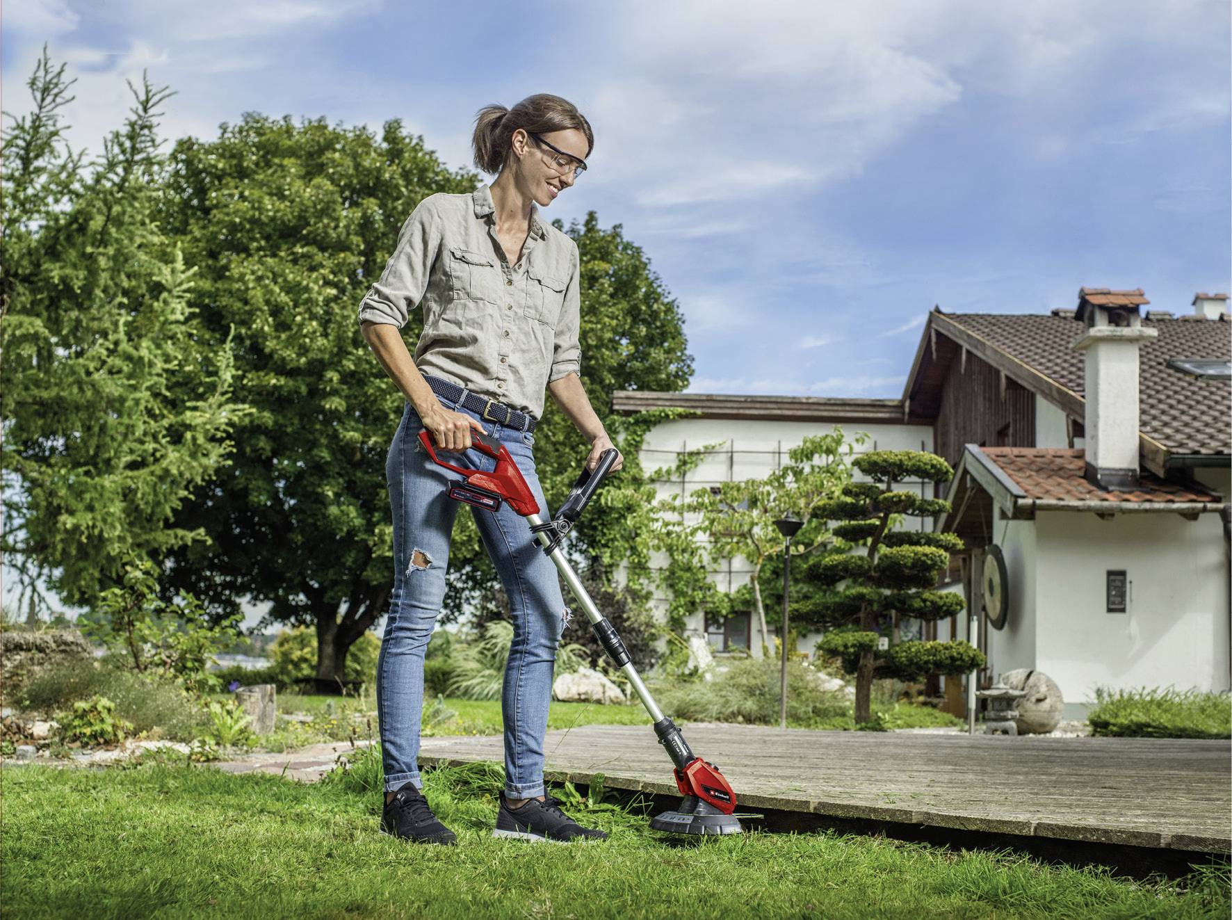 A woman is using a lawn trimmer in a garden next to a house. She is wearing jeans and a shirt. Trees and a blue sky are visible in the background.
