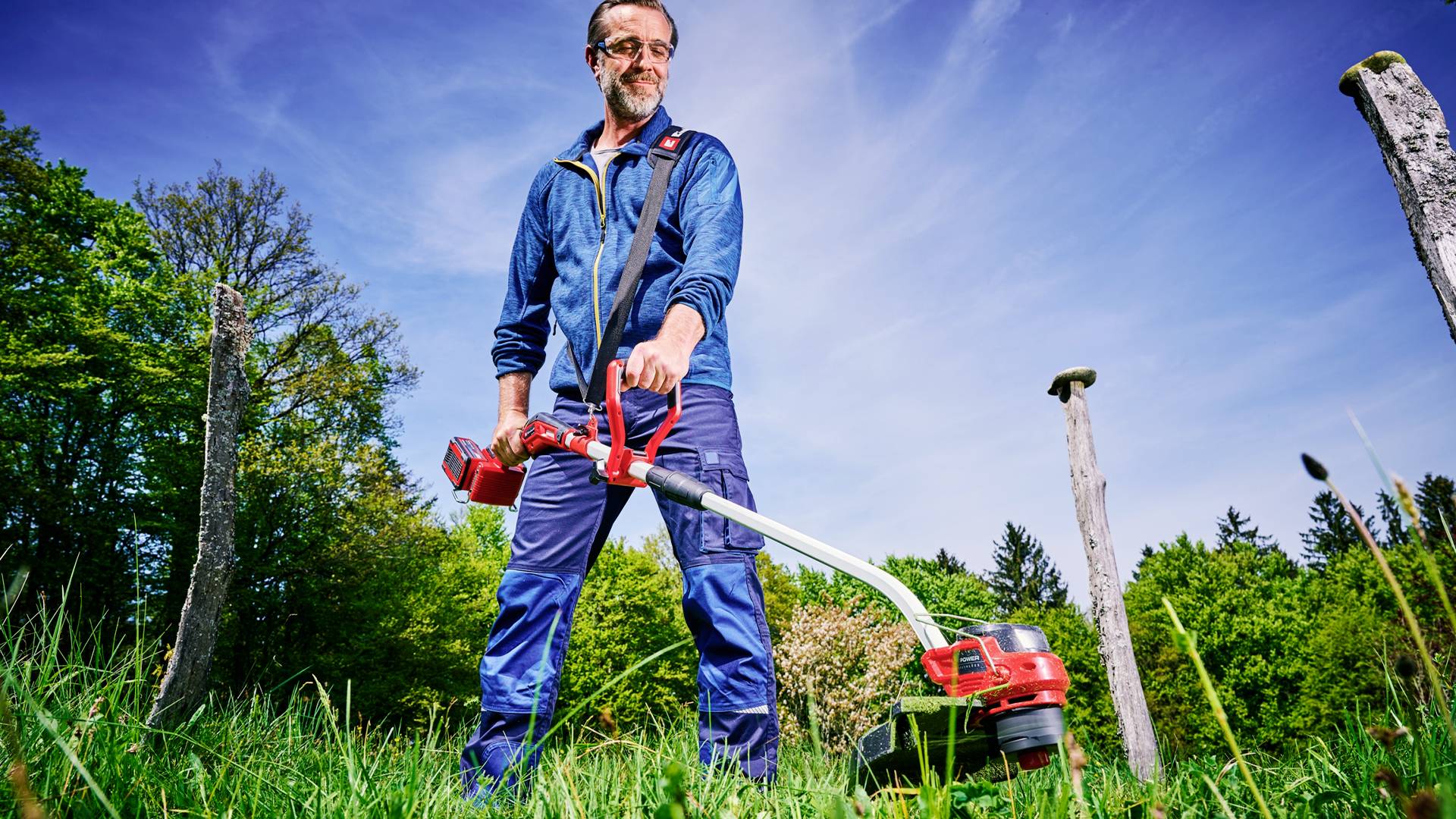A man is operating a red lawn trimmer outdoors, surrounded by trees and a blue sky.