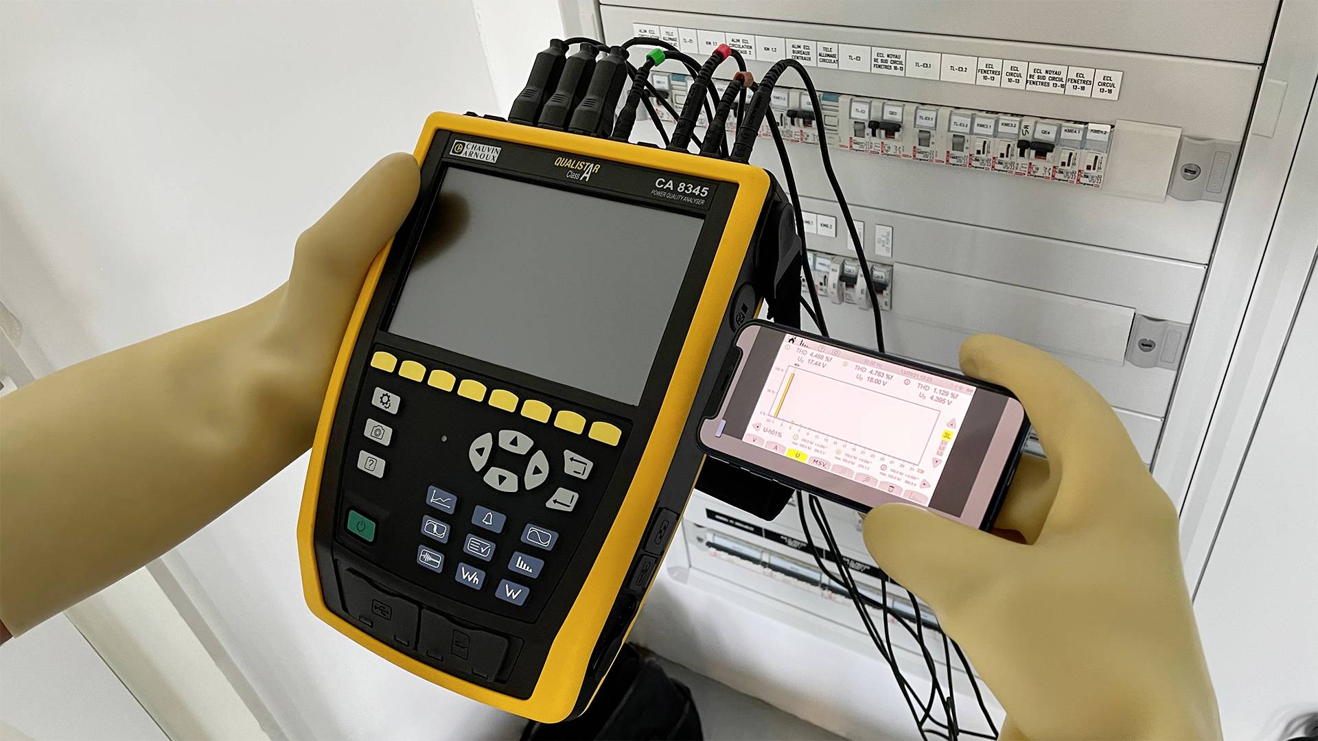 A technician is measuring electrical parameters with a portable measuring device and a smartphone in front of an open electrical cabinet.