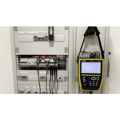 An open electrical cabinet with various switches and cables. On the right, a measuring instrument with a digital display hangs inside the cabinet.