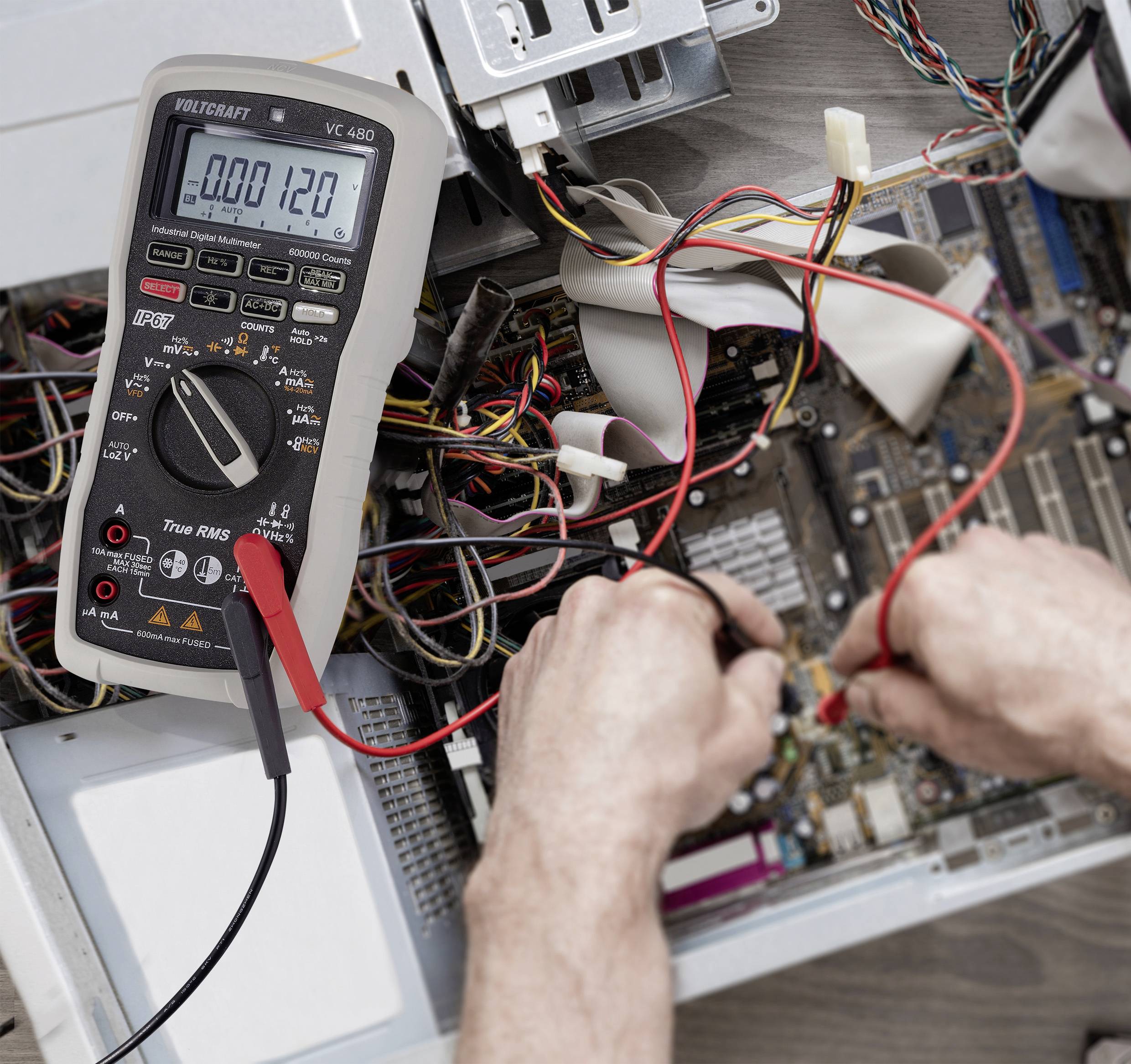 A person is measuring the current flow on an open computer circuit board with a multimetre. Many cables are visible.