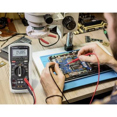 A person is repairing an electronic circuit board under a microscope, using a multimeter to measure voltage.