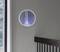 A round, contemporary lamp with blue light hangs on a grey wall. To the left, there is a window with a view of greenery.
