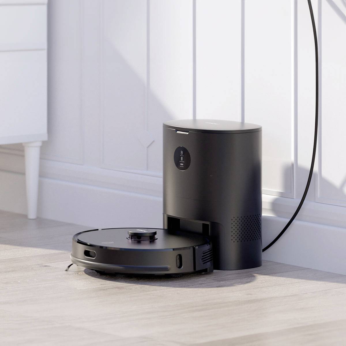 A black robotic vacuum cleaner docks at a charging station on a light-coloured room wall. In the background, part of a white desk is visible.
