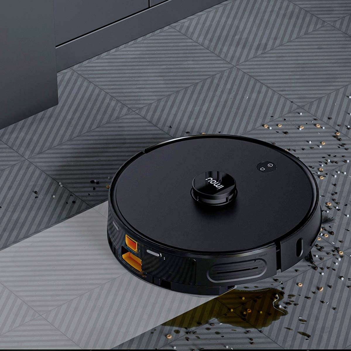 A black robotic vacuum cleaner is cleaning a tiled floor by sucking up small crumbs. Kitchen cupboards are visible in the background.