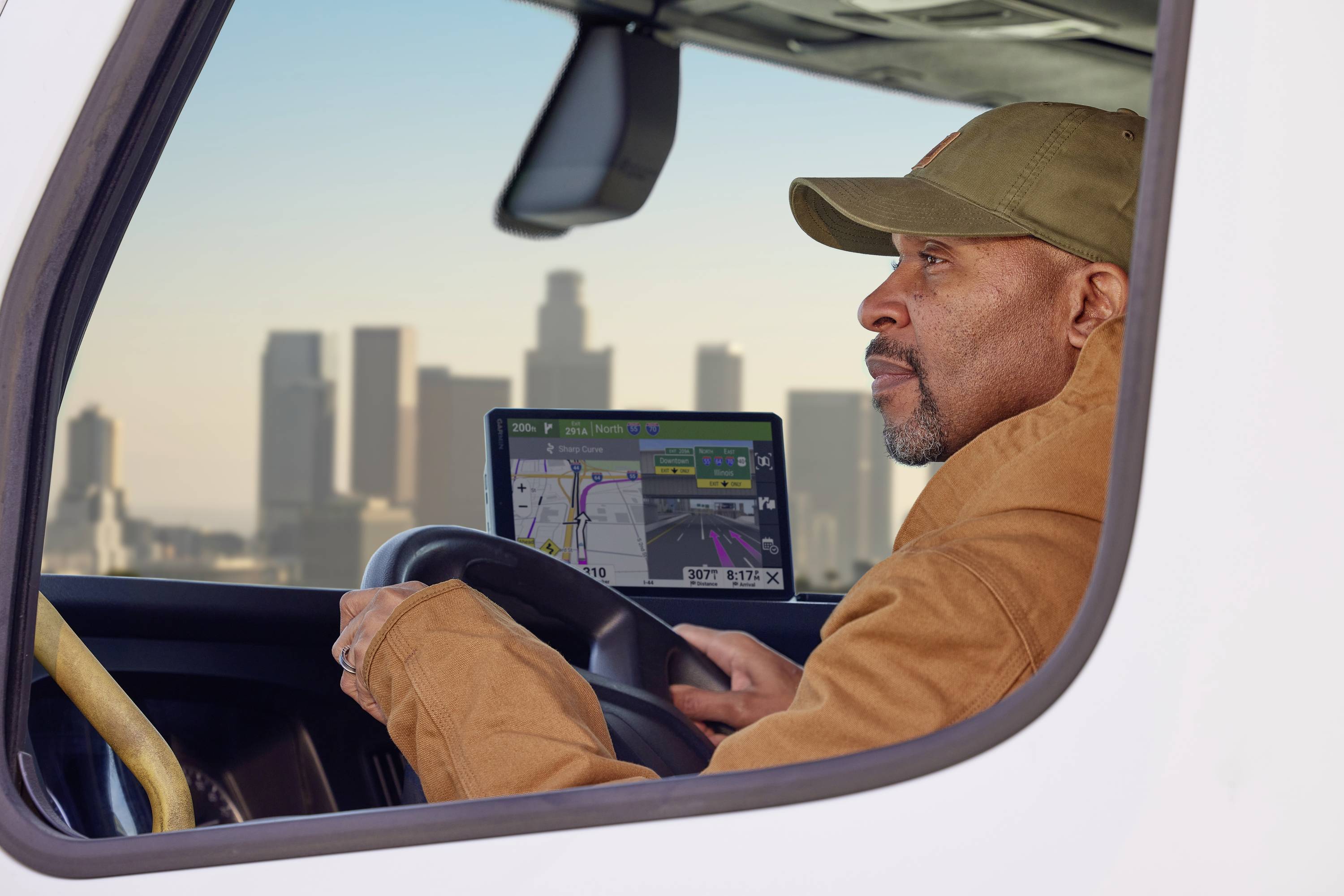 A man sits in the cab of a lorry, looking intently outside. A cityscape is visible in the background.