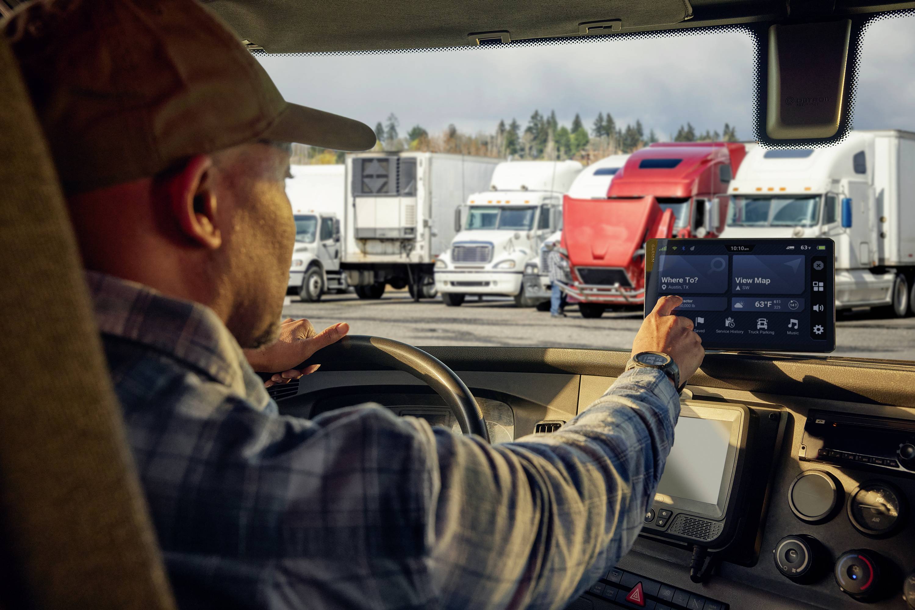 Lorry driver tapping on a satellite navigation device in the cab, while standing at a motorway service station with parked lorries in the background.