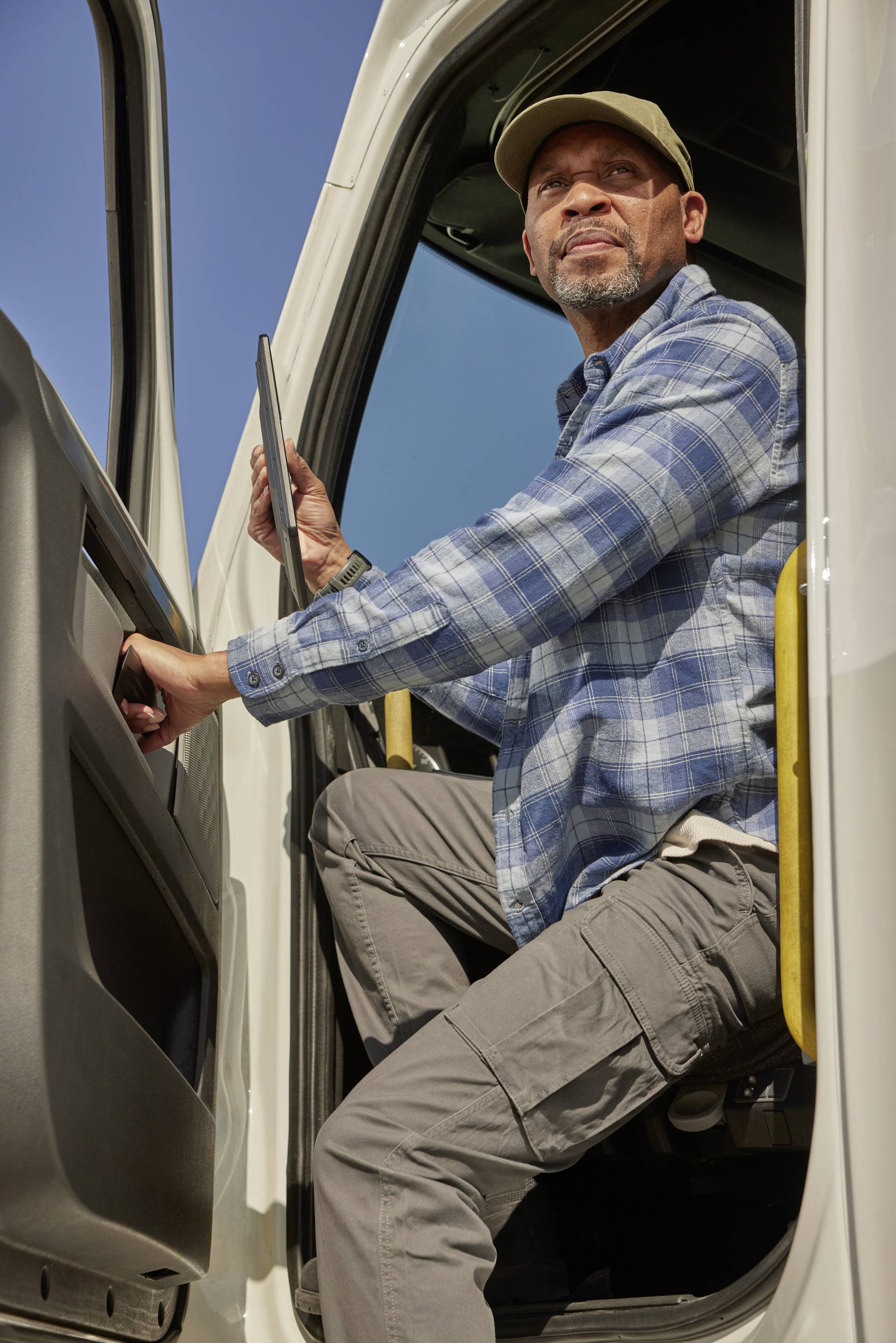 A man in a checked shirt and cap sits in an open lorry, looking attentively ahead and holding a tablet.