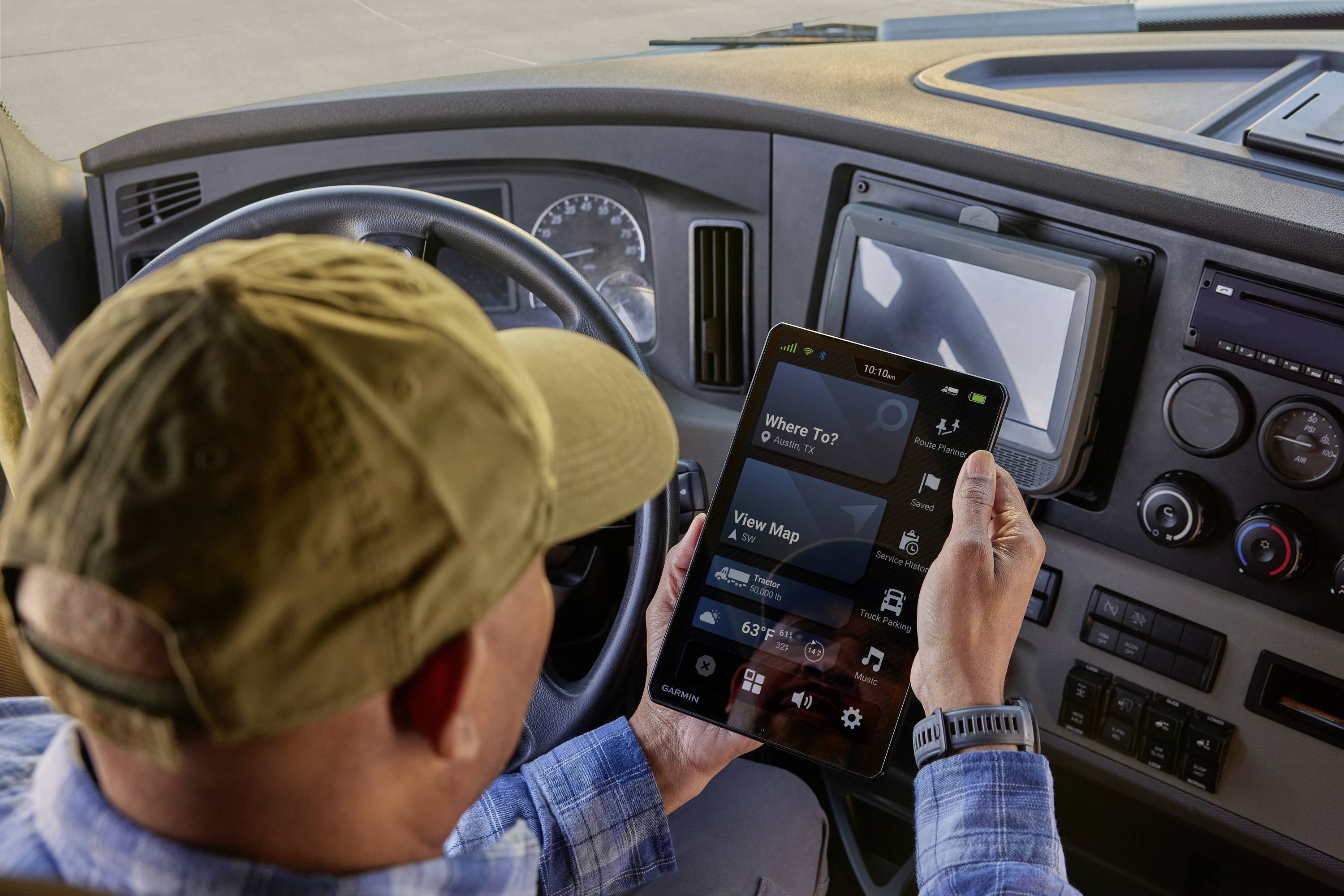 A driver sits in a lorry and holds a tablet displaying a navigation app. The dashboard is visible in the background.