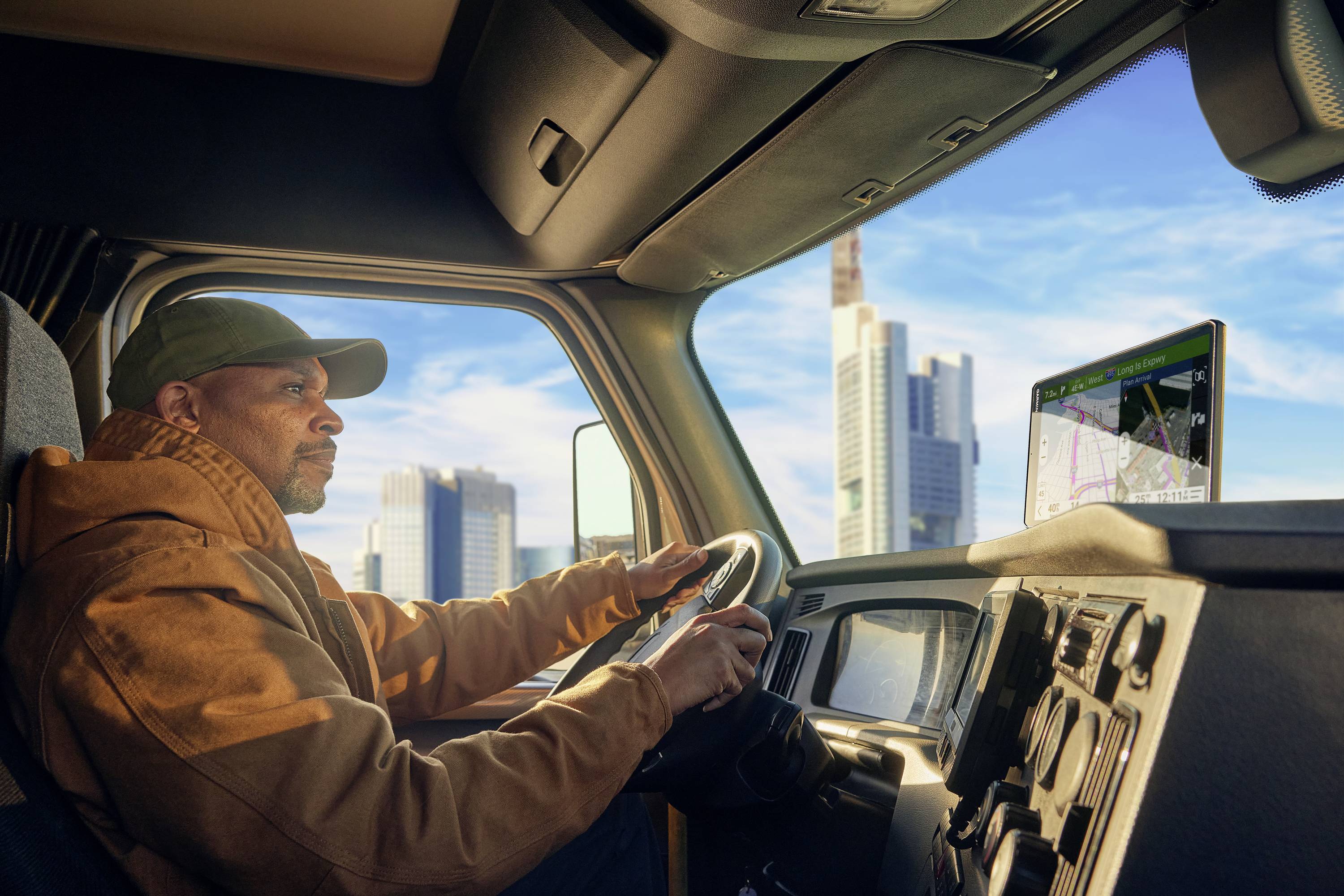 A man in a brown jacket is driving a lorry through a city with tall buildings. A satellite navigation system is displaying a map.