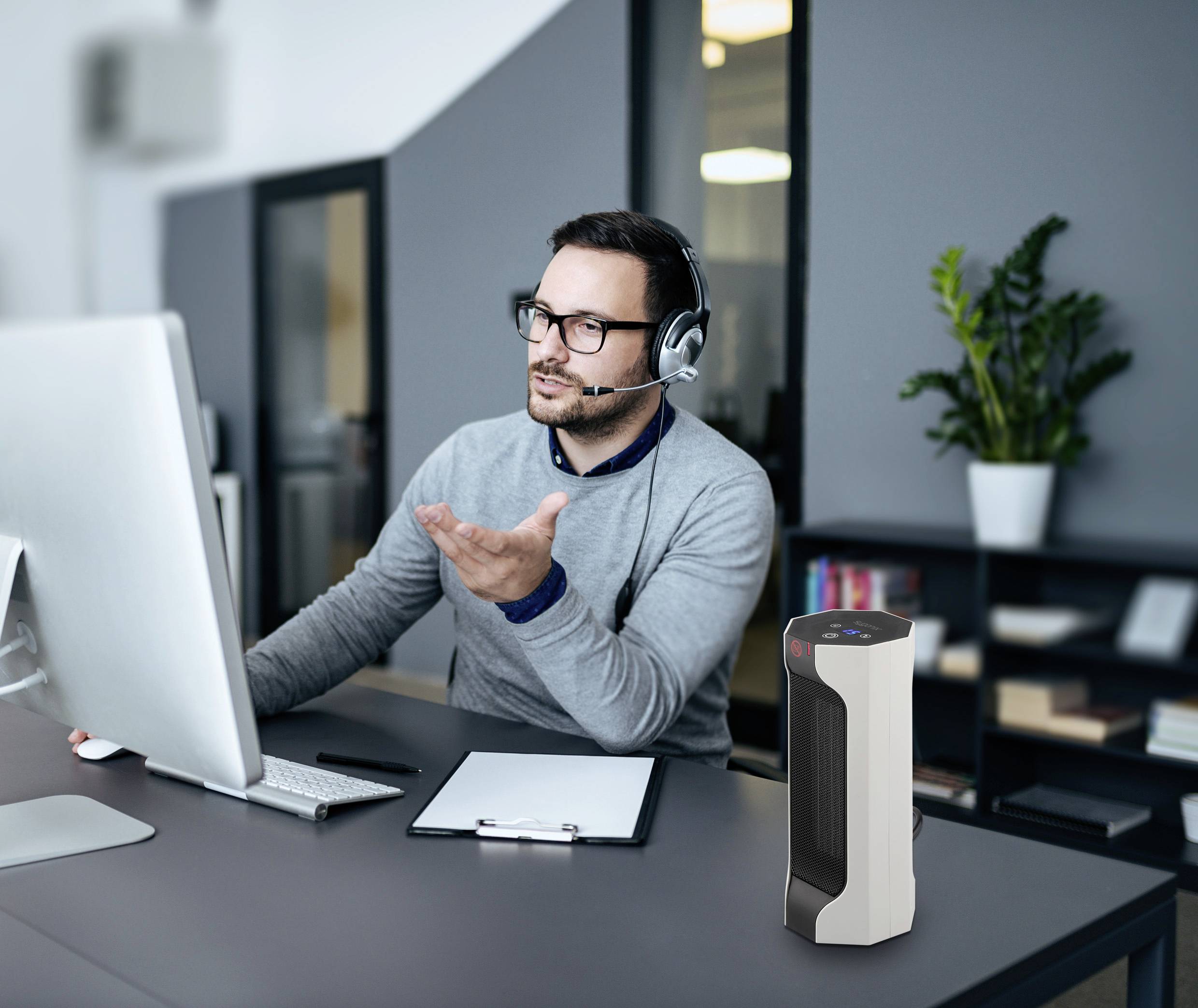 A man wearing headphones is working at a desk with a computer and notepad. A plant stands in the background.
