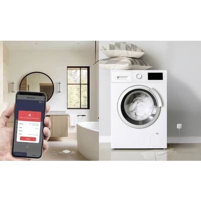 A hand holds a smartphone with a water alarm app, next to a washing machine with water puddles on the floor.