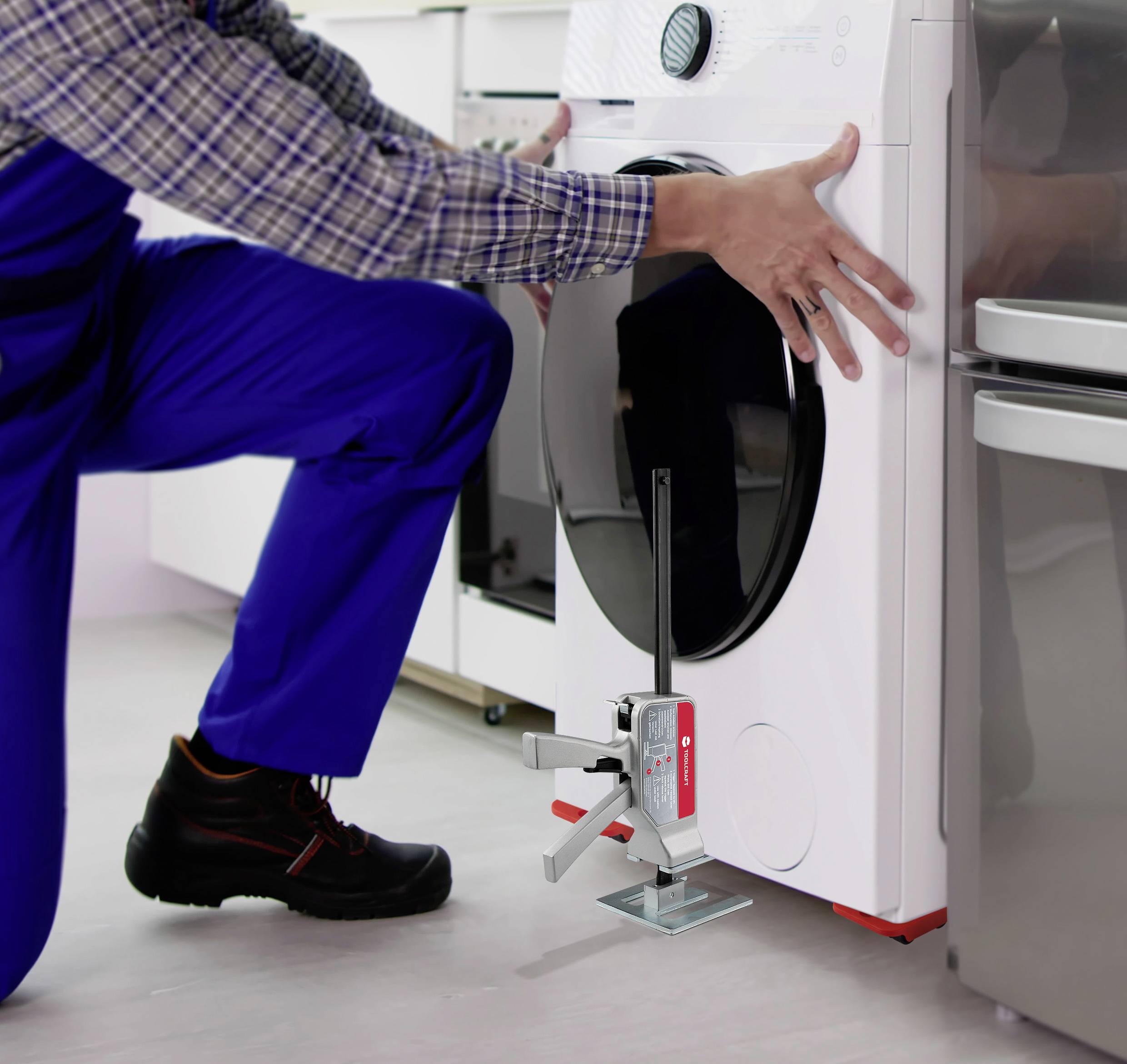 A person in blue workwear is levelling a washing machine with an installation tool in a modern utility room.