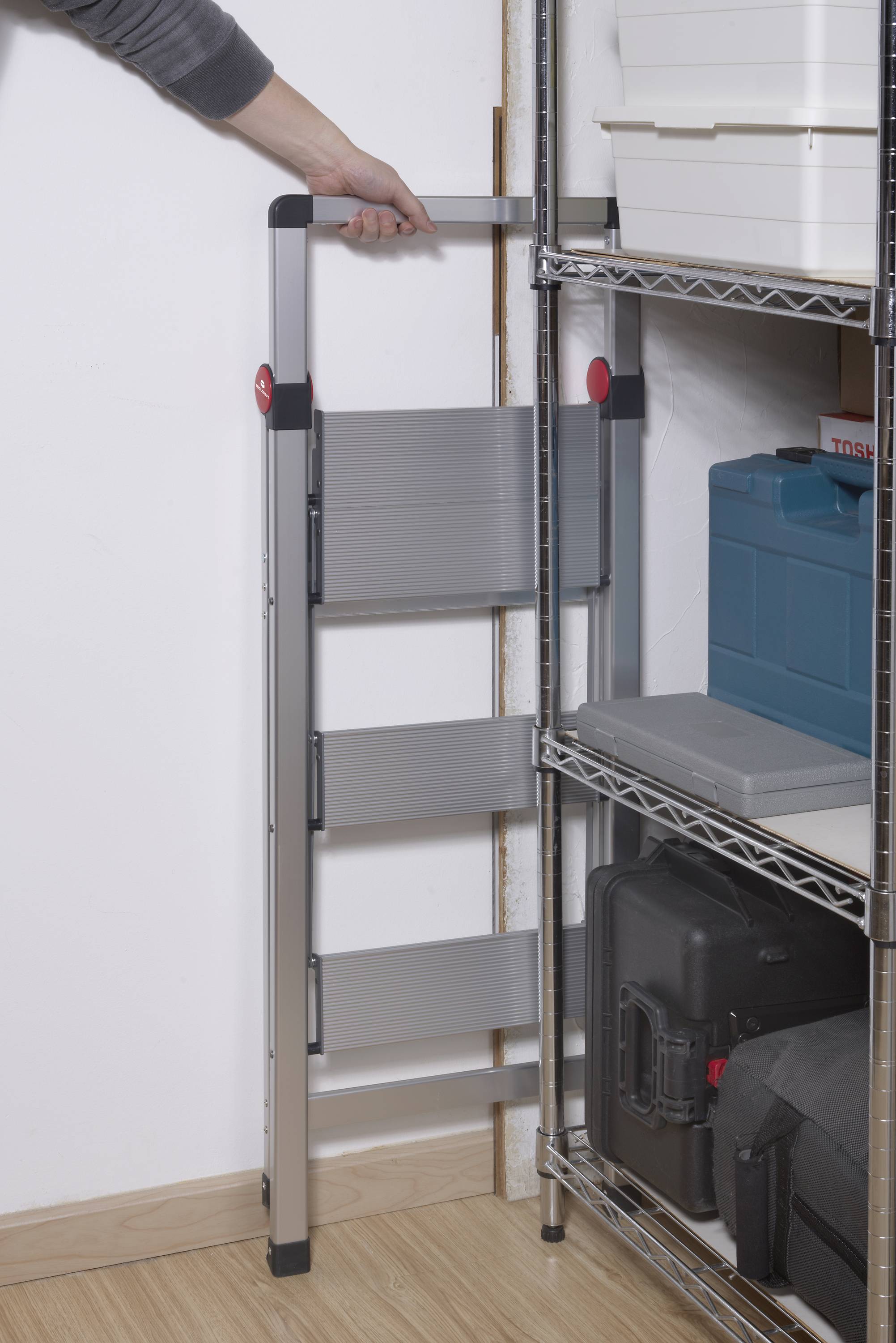 A person is pulling out a compact folding ladder from a narrow gap between a shelf and a wall.