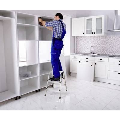 A man in work attire is standing on a ladder and assembling a piece of furniture in a white kitchen.