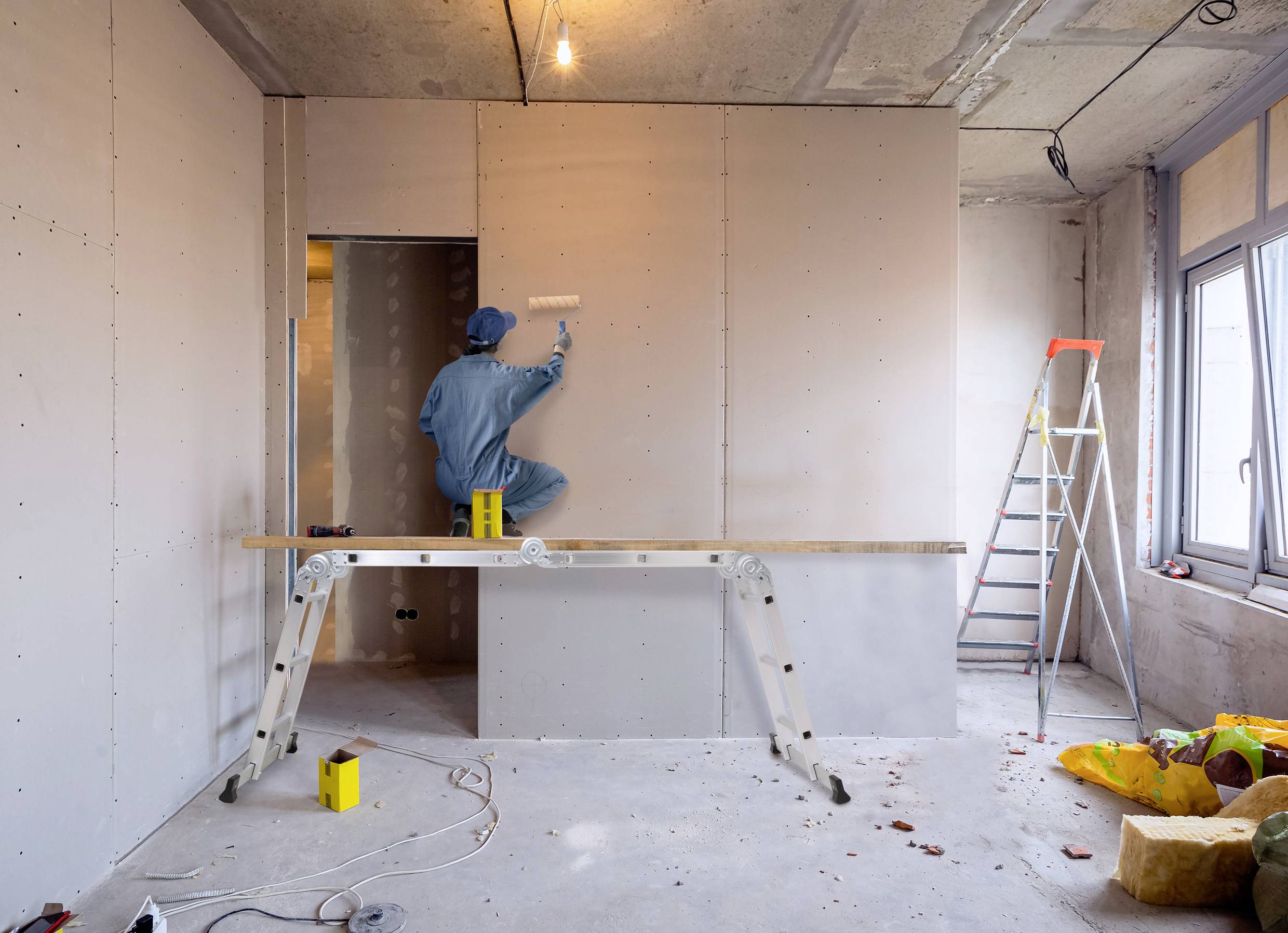 A construction worker in blue overalls is painting a plasterboard wall in a room with ladders and building materials.
