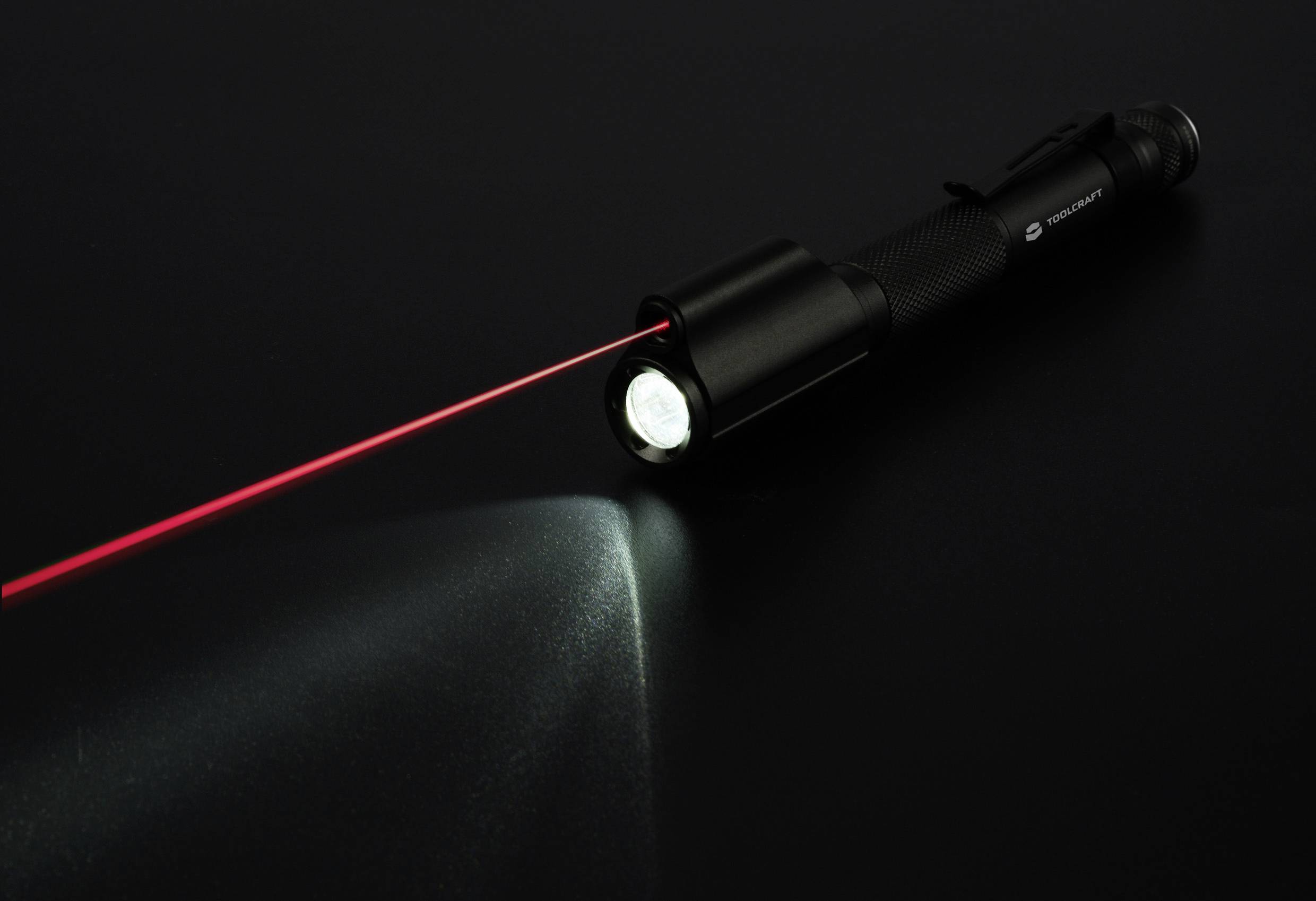A black torch on a dark surface emits a red laser beam. The light cone is slightly visible.