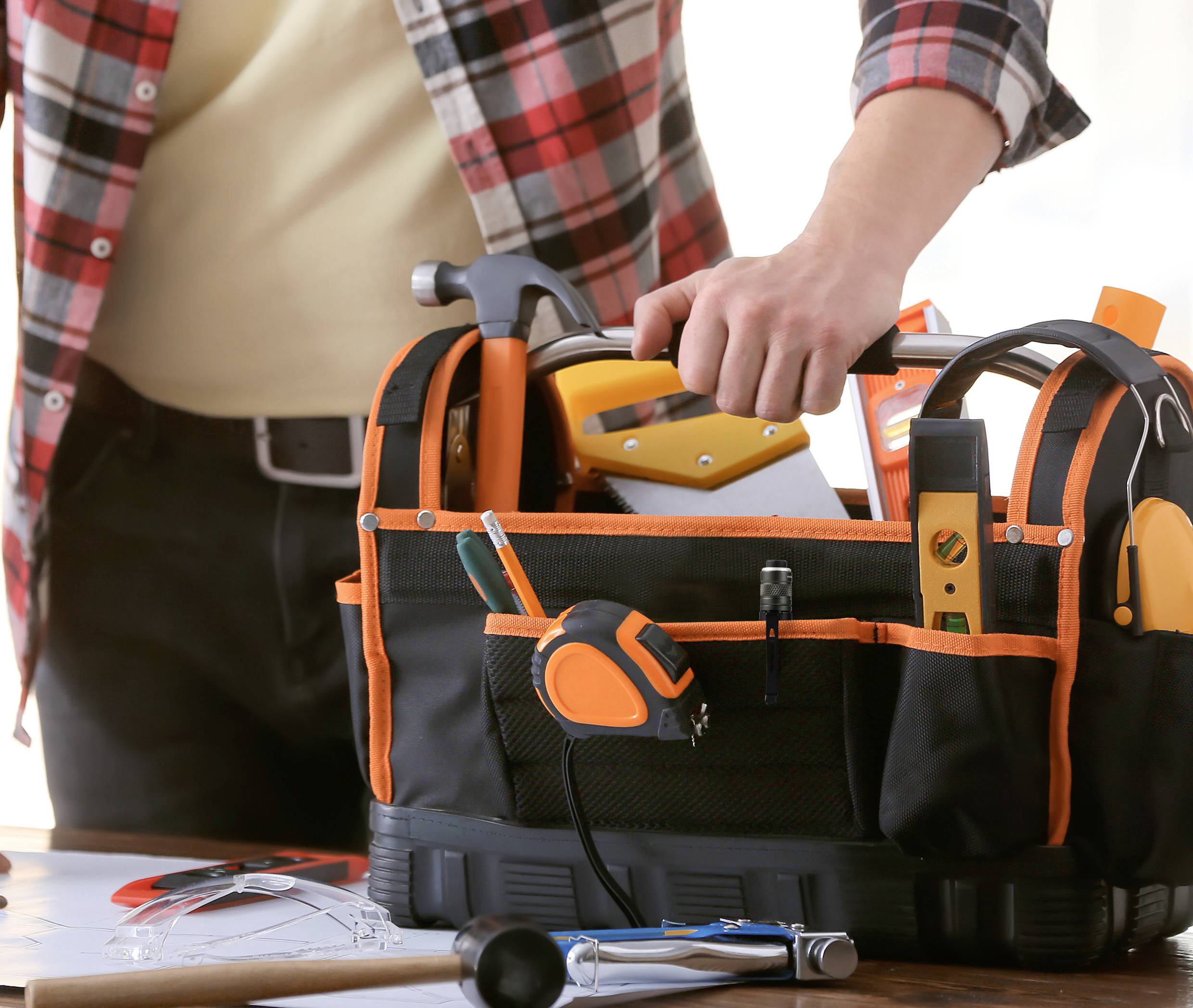 A person is packing a tool bag with various hand tools on a table covered with drawings and notes.