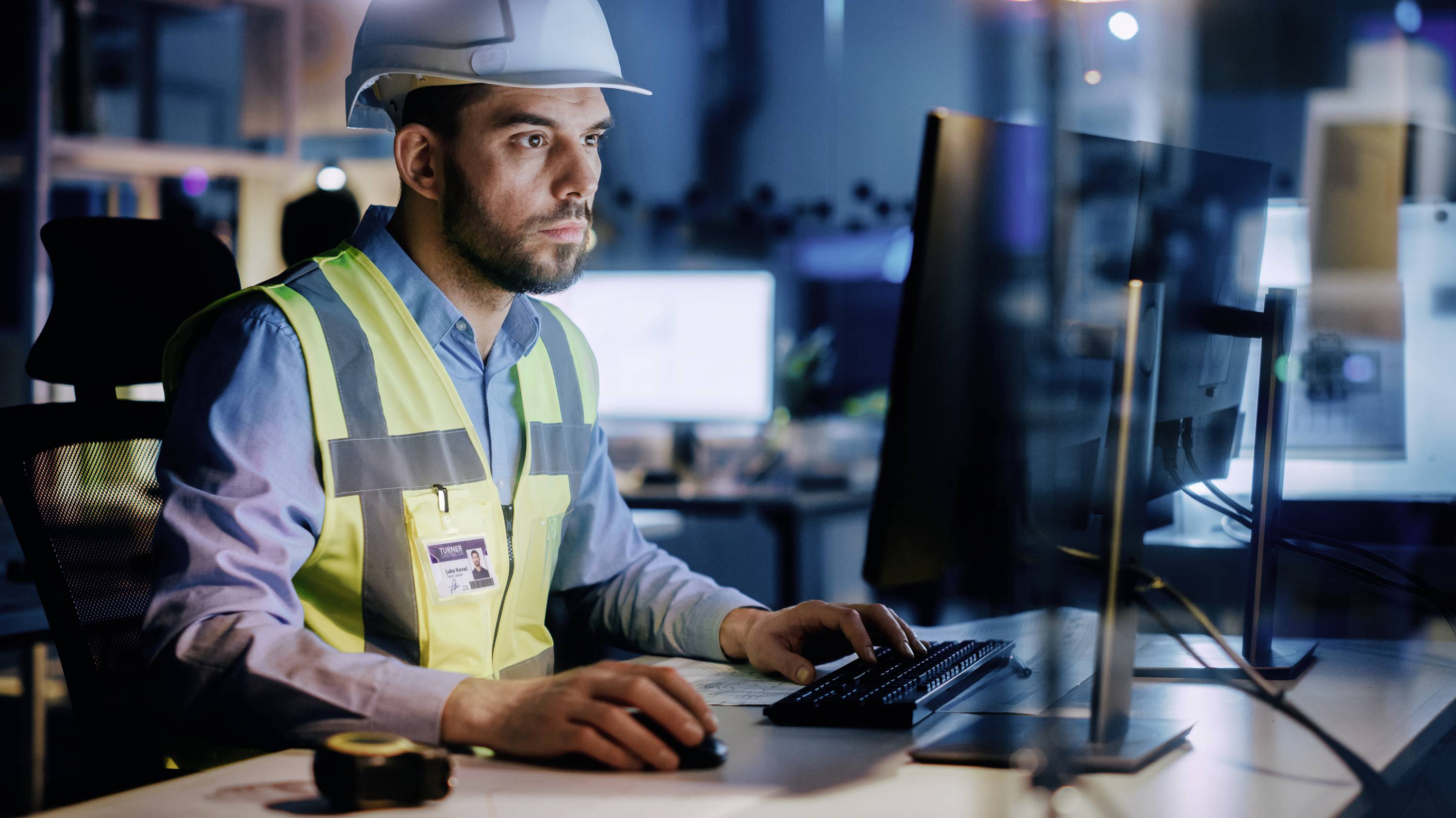 A man wearing a hard hat and high-visibility vest is working at a computer screen in an office, possibly in a construction-related environment.