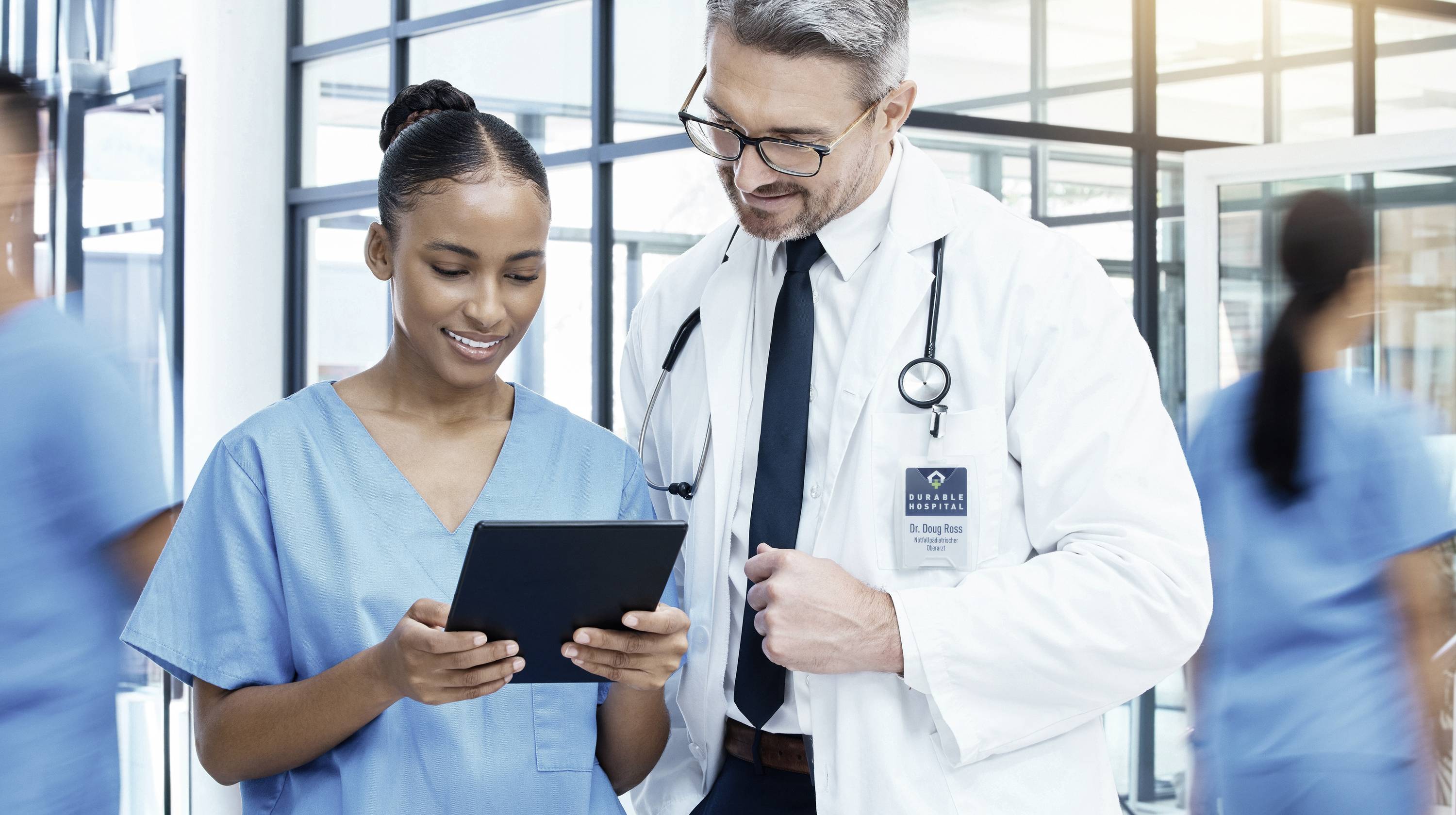 A doctor and a nurse smile and look together at a tablet in a hospital corridor.