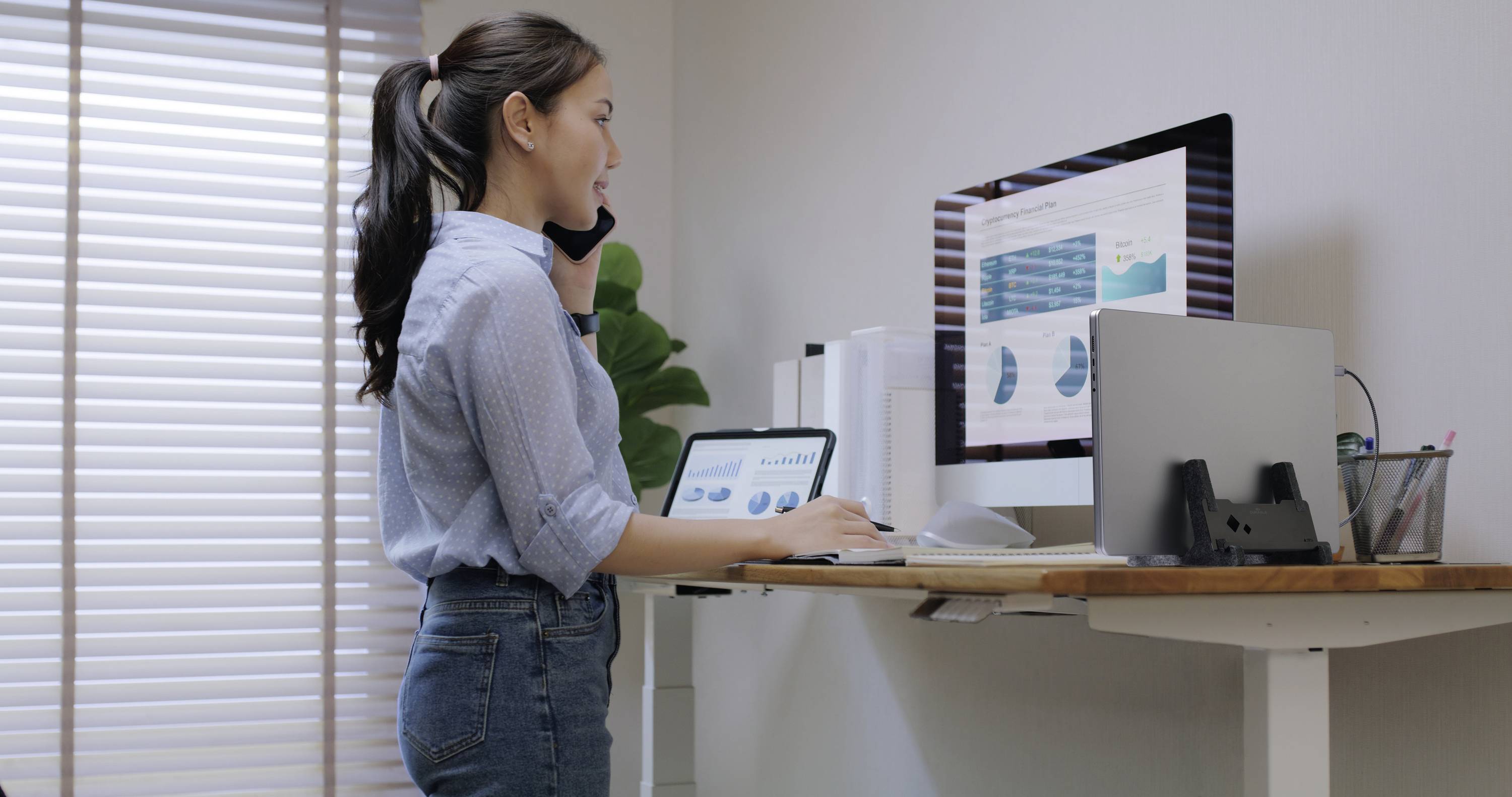 A woman is standing at a standing desk, on the phone and examining diagrams on a computer screen, with a tablet beside her.