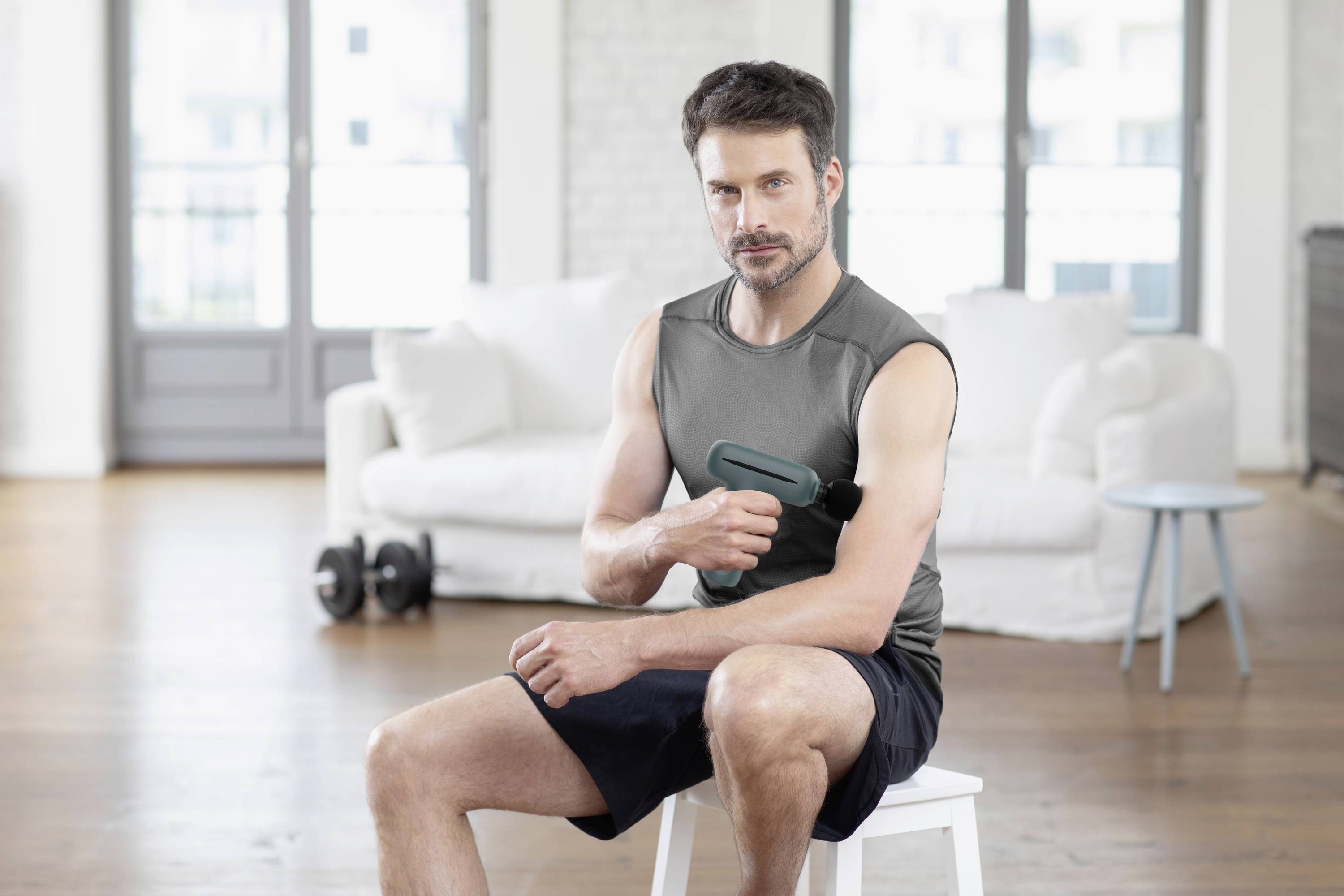 A man is using a massage device on his upper arm, sitting on a stool in a bright room with dumbbells in the background.