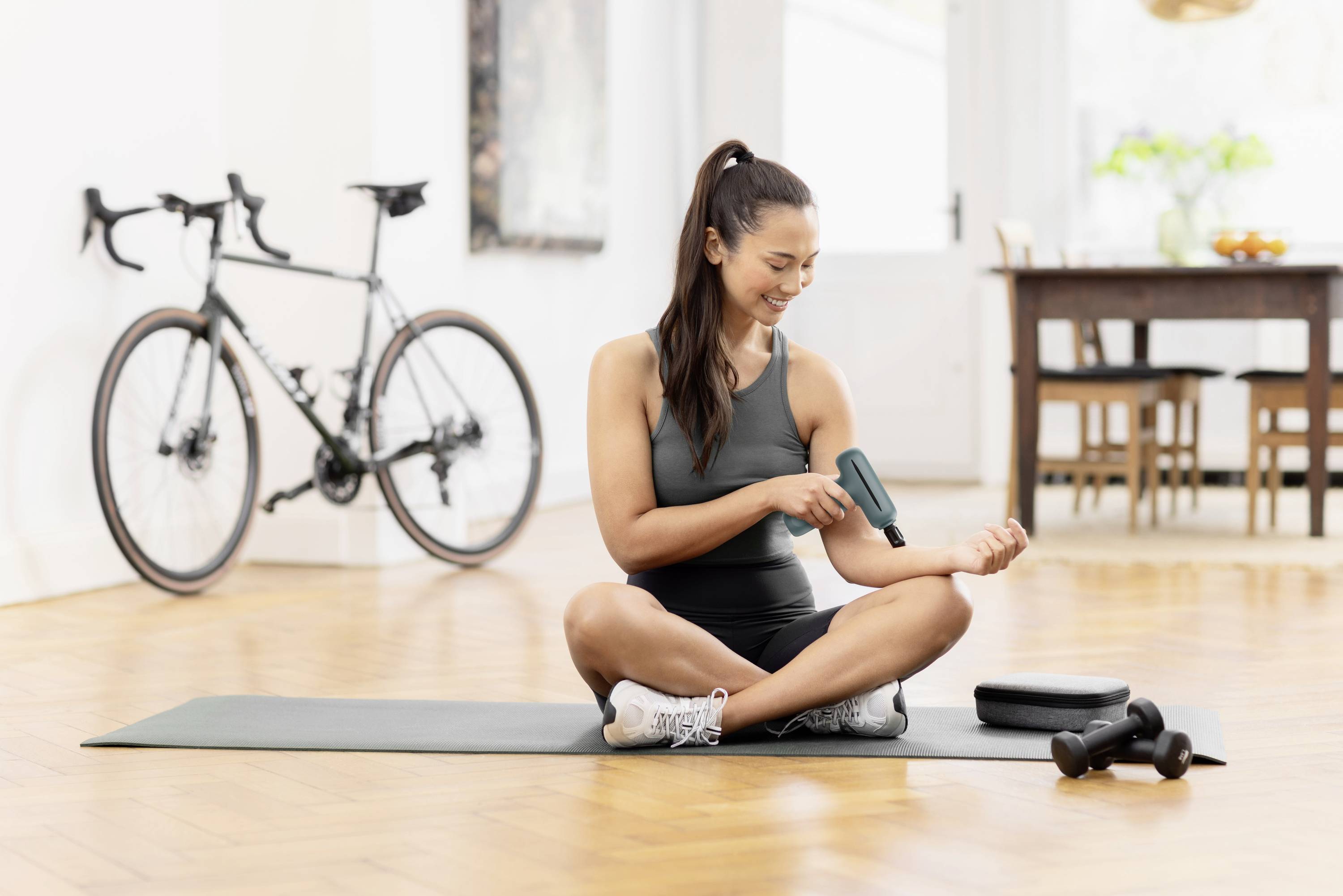 A woman sits on a yoga mat, using a massage gun on her arm. A bicycle is in the background, with dumbbells on the floor.