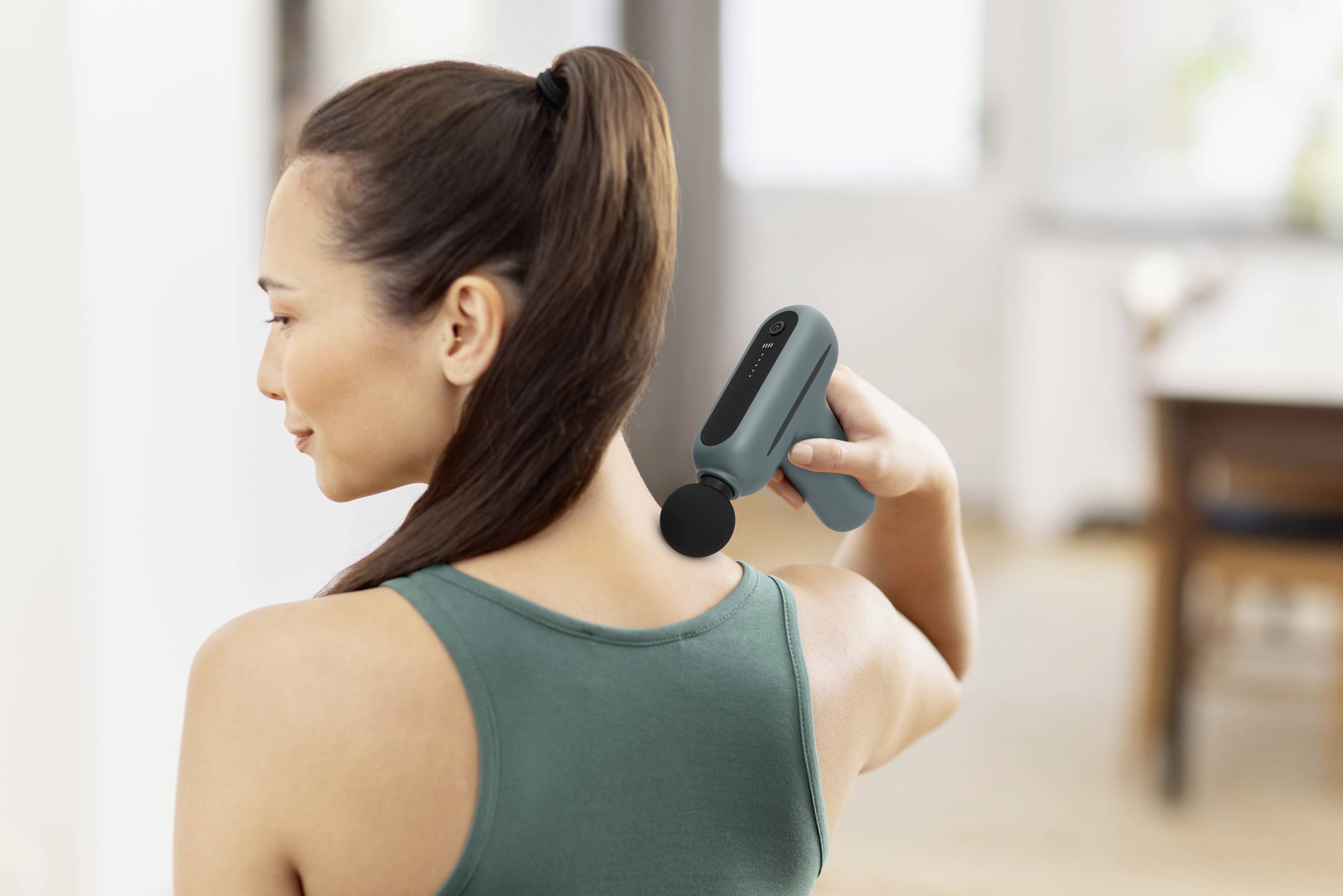 A woman in a green top is using a massage device on her neck. She is standing in a bright room.