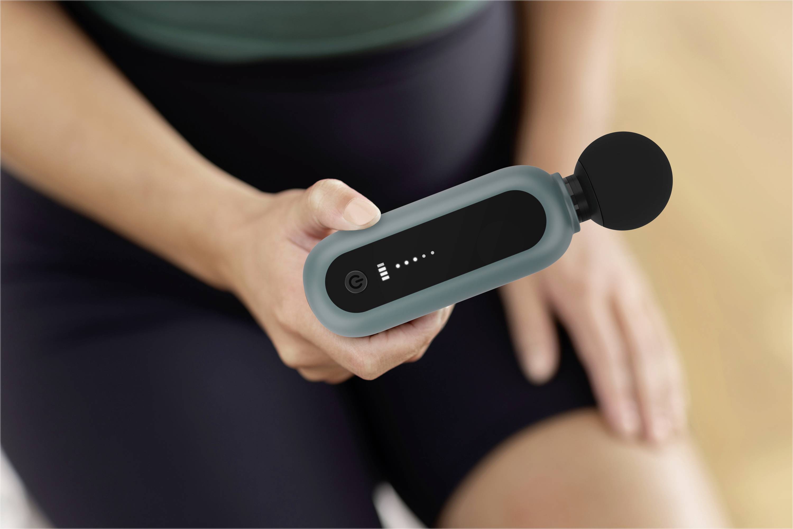A person is holding a hand massage device with five illuminated indicators showing the battery level. Their other hand is resting on their thigh.