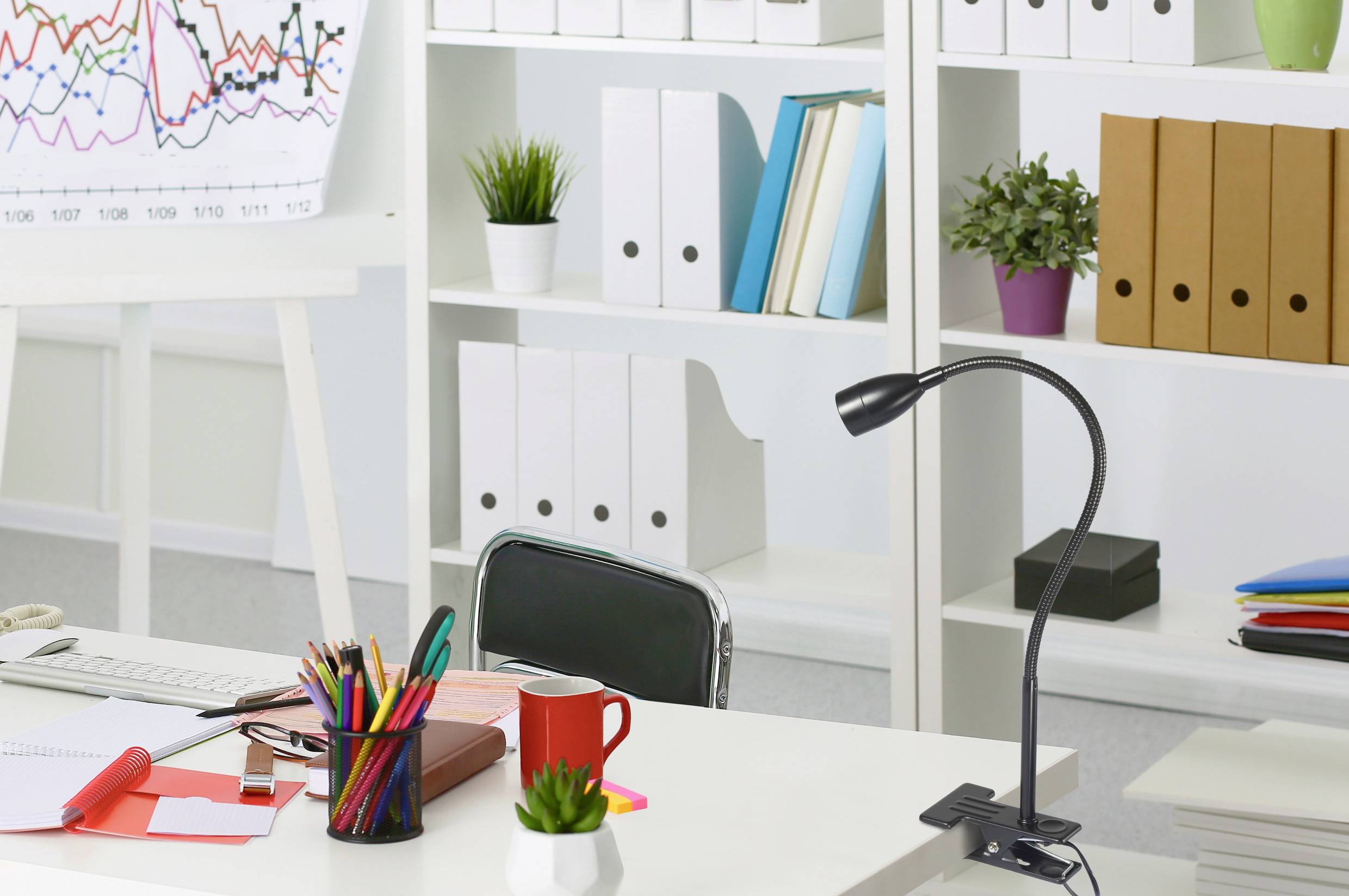 Office with a desk, lamp, mug, writing utensils, and laptop in front of a bookshelf with folders and plants. Flipchart in the background.