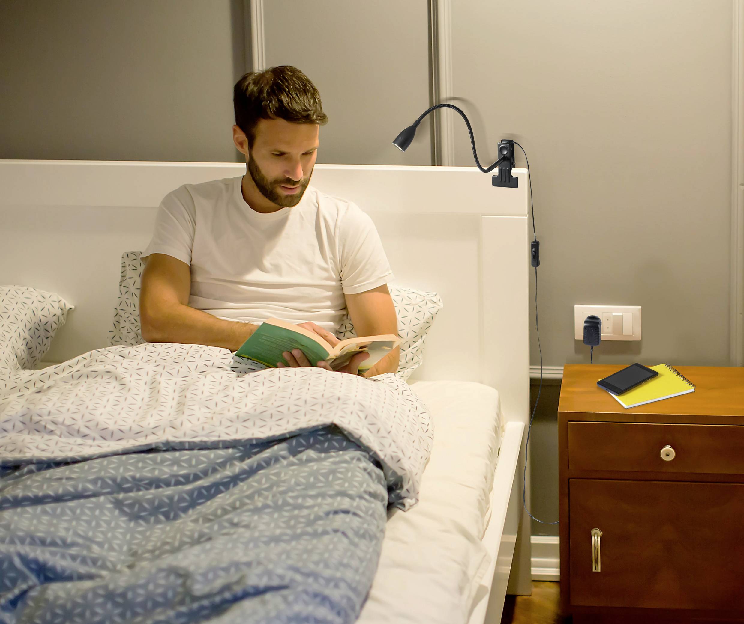 A man is sitting in bed reading a book. A smartphone and a notebook are lying next to him on the bedside table.