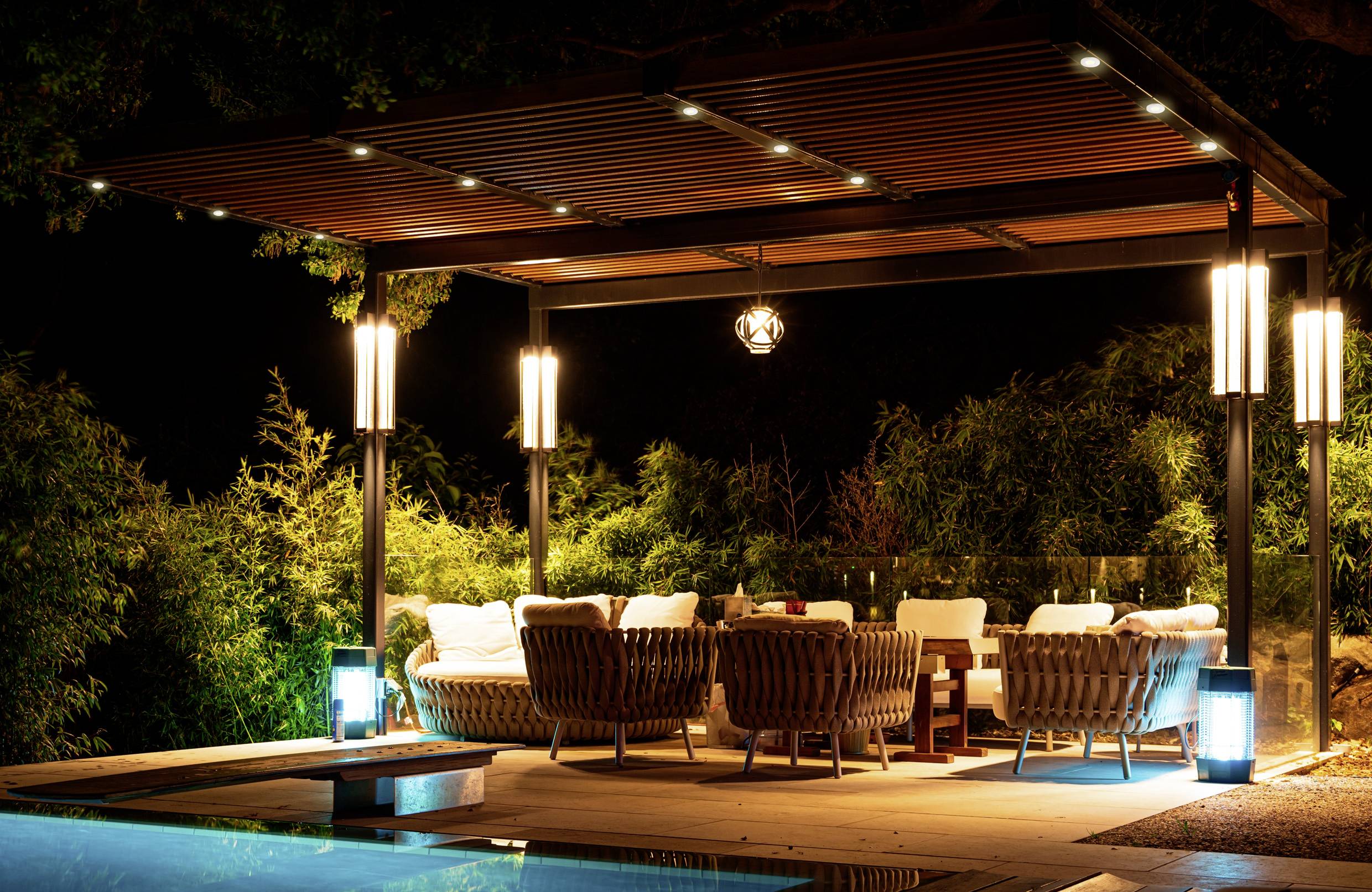 A well-lit outdoor lounge at night with cozy wicker chairs and a wooden pergola, situated beside a pool, surrounded by lush greenery.