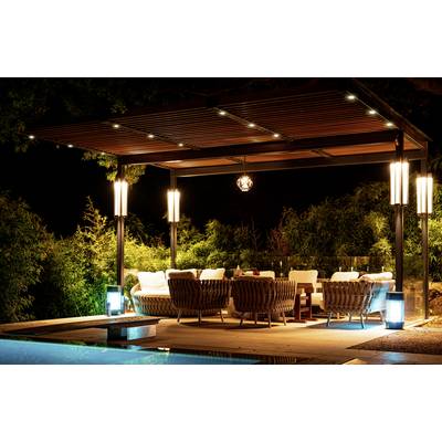 A well-lit outdoor lounge at night with cozy wicker chairs and a wooden pergola, situated beside a pool, surrounded by lush greenery.