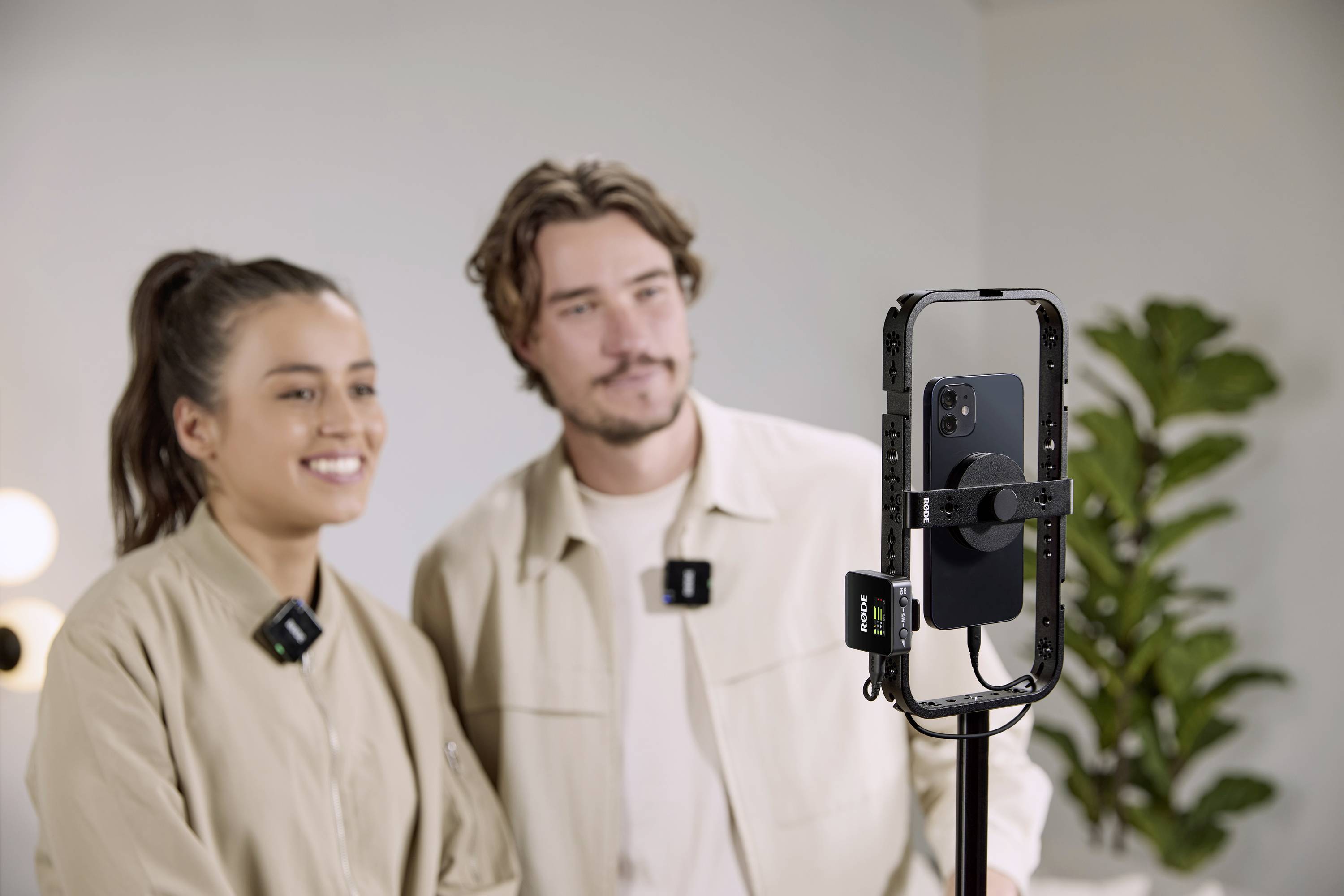 Two people are standing smiling in front of a smartphone mounted on a tripod. A plant is visible in the background.