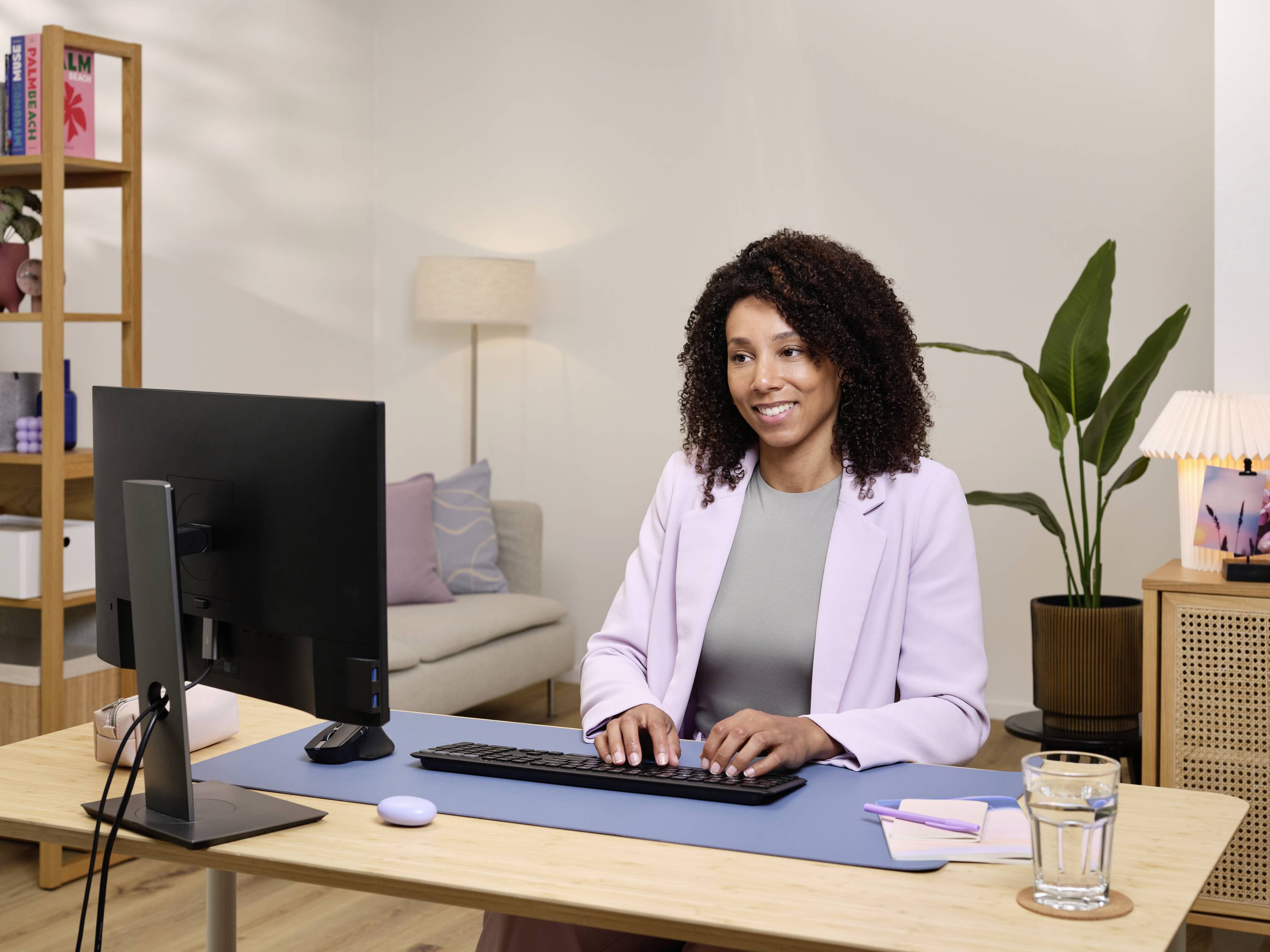 A woman sits at a desk in front of a computer in a modern office. Plants and lamps are visible in the background.