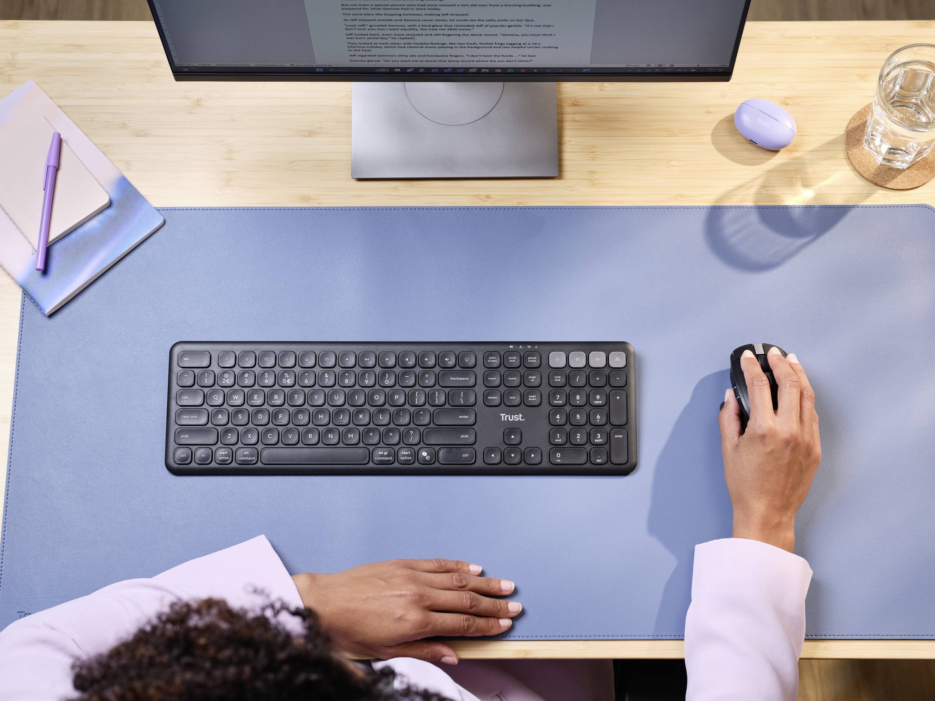 A person is working at a desk with a keyboard and mouse on a blue mat in front of a monitor. Office supplies and a drink are visible.