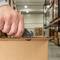 A person opens a cardboard box with a utility knife in a warehouse. Shelves with additional boxes can be seen in the background.