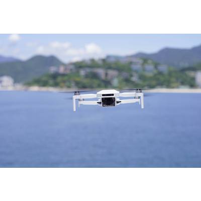 A white drone hovers over blue water with blurred green hills in the background.