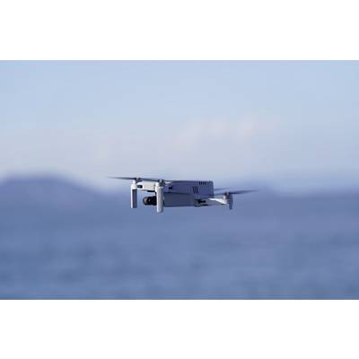 A white drone hovers above the blue sea. Blurred hills are visible in the background.