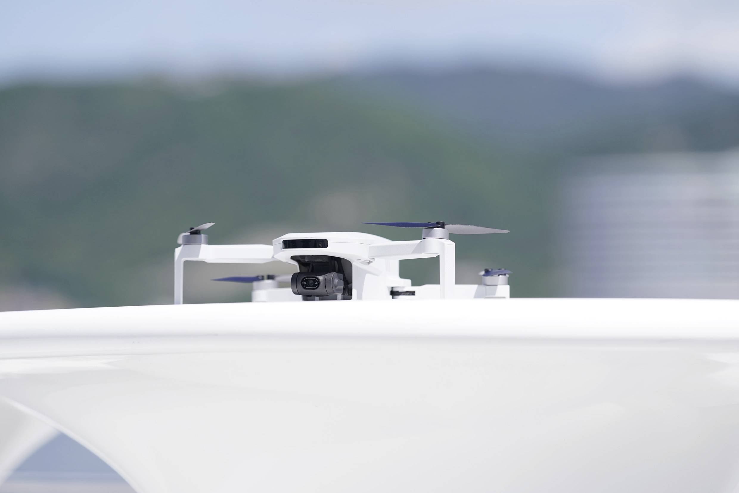 A white drone with a camera hovers above a smooth, light surface. Blurred trees and a sky are visible in the background.