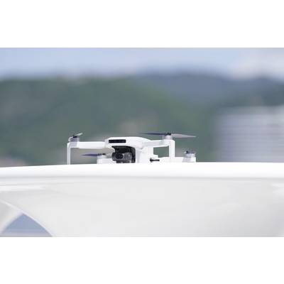 A white drone with a camera hovers above a smooth, light surface. Blurred trees and a sky are visible in the background.