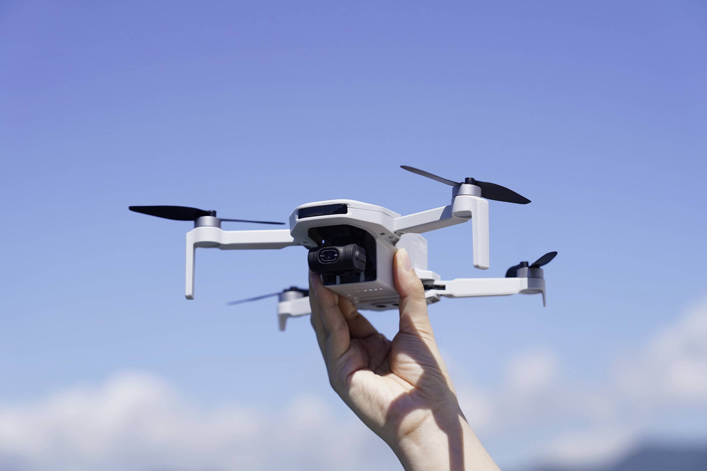 A hand holds a small drone against a blue sky. The drone has four propellers and a camera.
