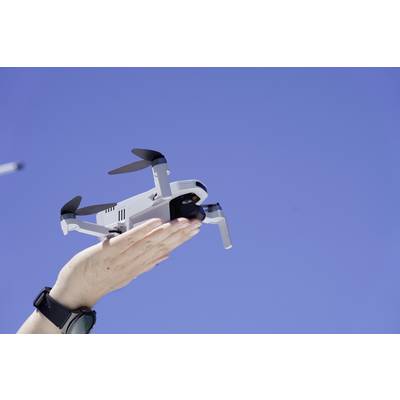 A hand holds a small drone beneath a clear, blue sky. The drone is ready for takeoff and the person is wearing a wristwatch.