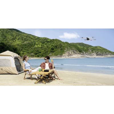 A couple sits relaxed on the beach in front of a tent, surrounded by green hills. A drone flies over the water in the background.