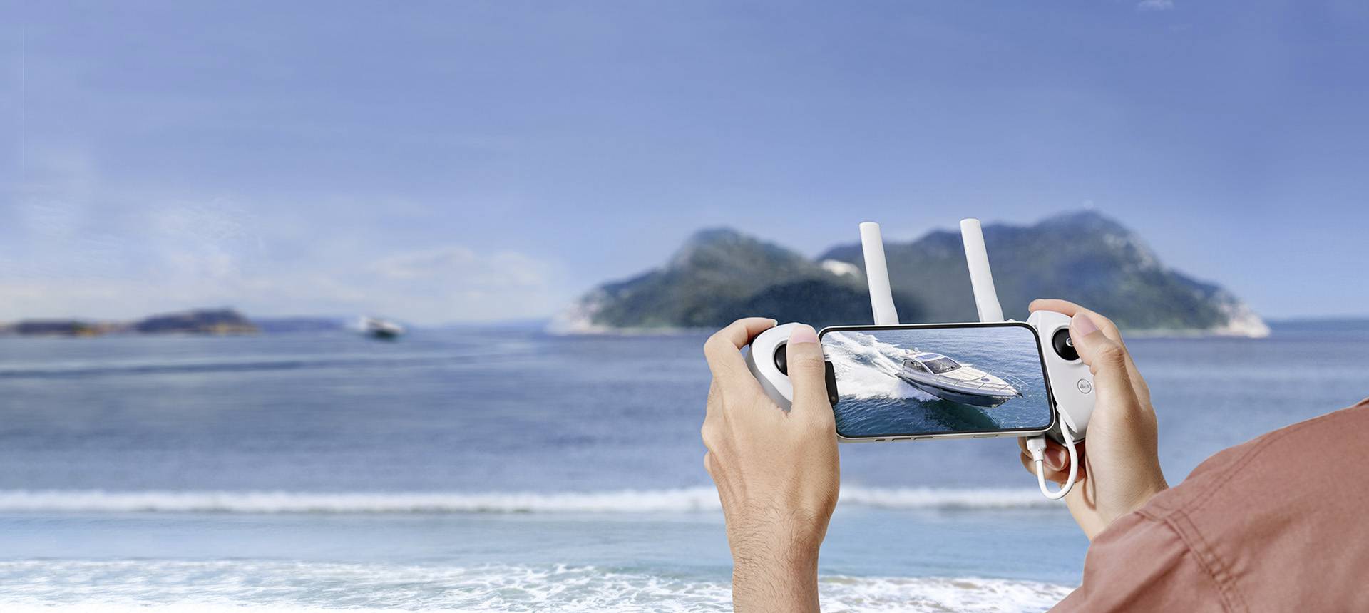 A person is holding a drone remote control displaying a catamaran on a smartphone screen. In the background, the sea and islands are visible.