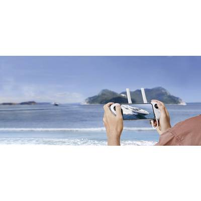 A person is holding a drone remote control displaying a catamaran on a smartphone screen. In the background, the sea and islands are visible.