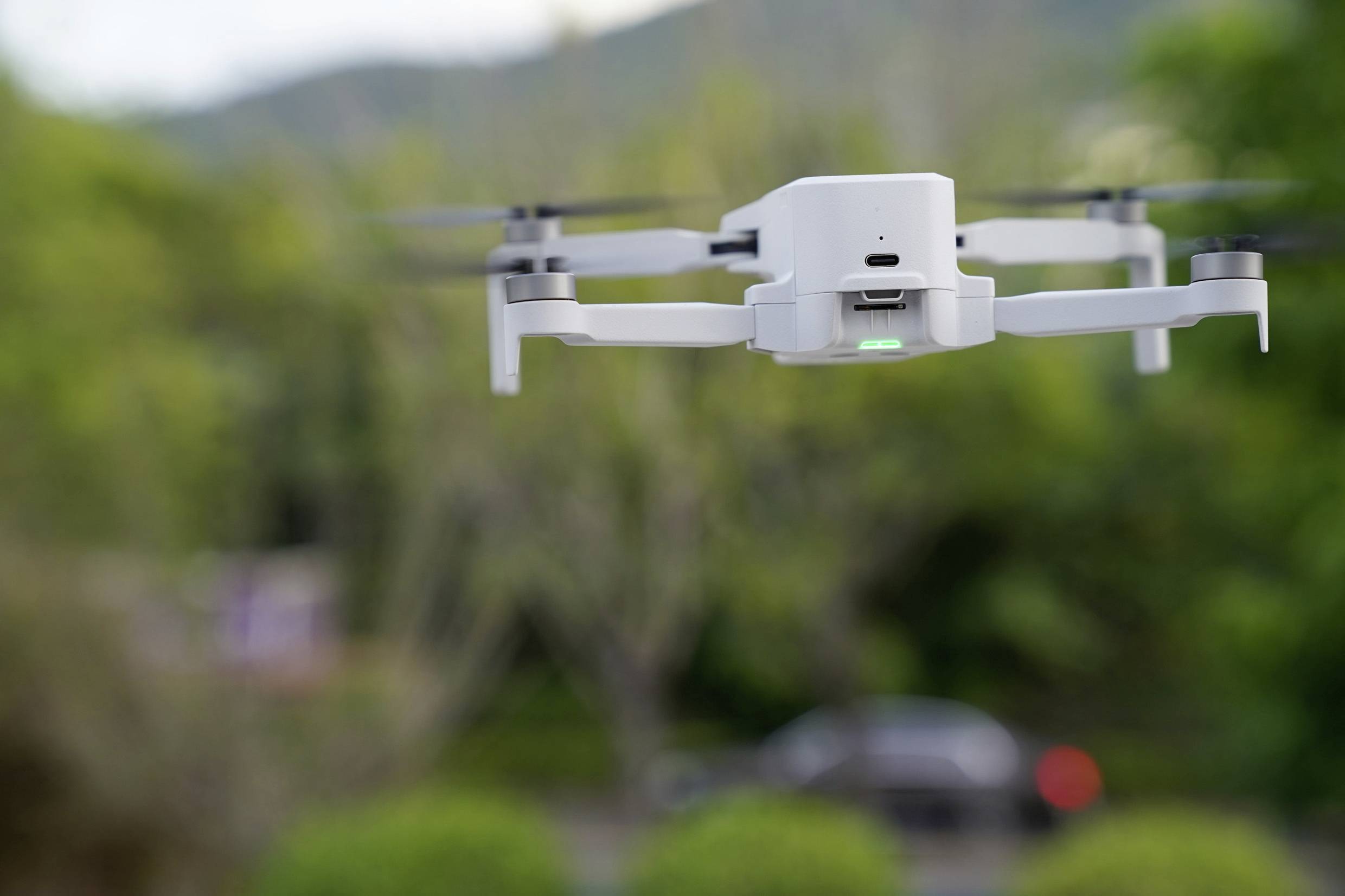 A white drone hovers outdoors in front of a blurred green landscape. It has four rotors and a bright green light on its front.